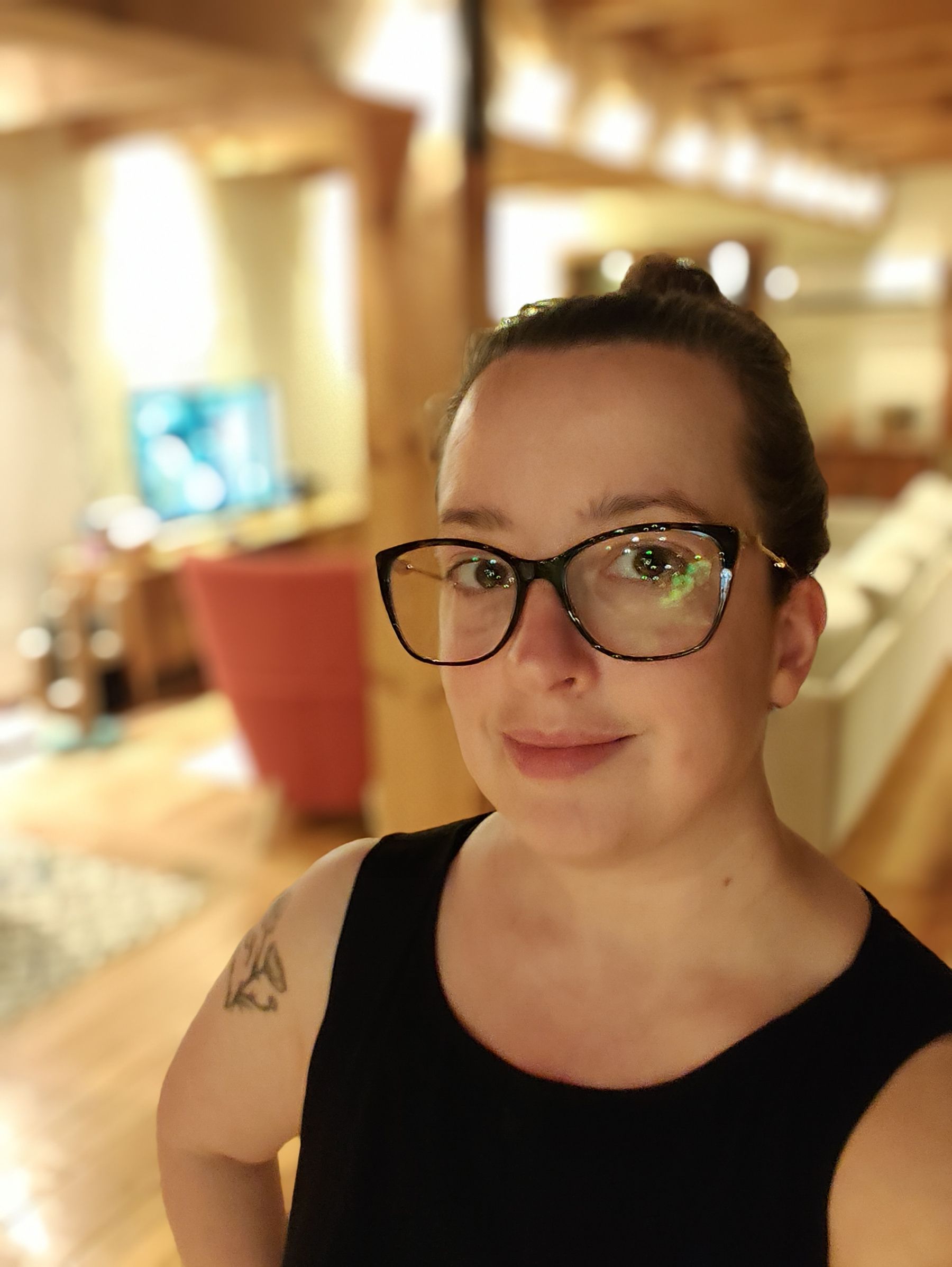 A woman wearing glasses and a black sleeveless top is smiling softly while standing in a warmly lit, cozy living room with wooden beams and furniture.