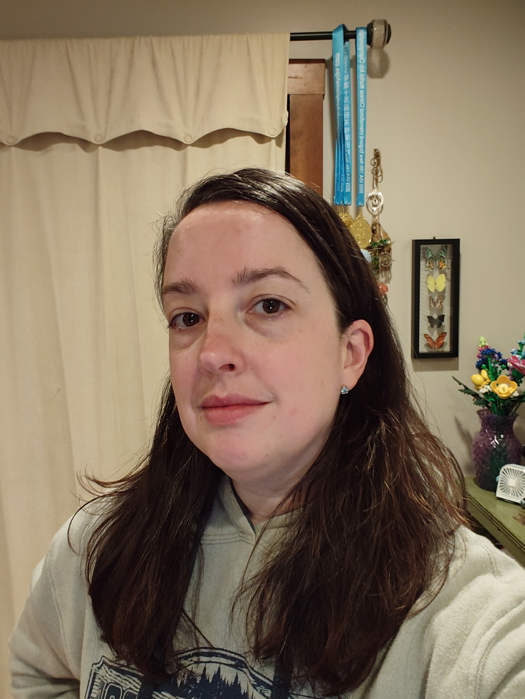 A woman with long brown hair is wearing a light-colored hoodie and standing indoors with decorations, including medals and a framed butterfly display, visible in the background.