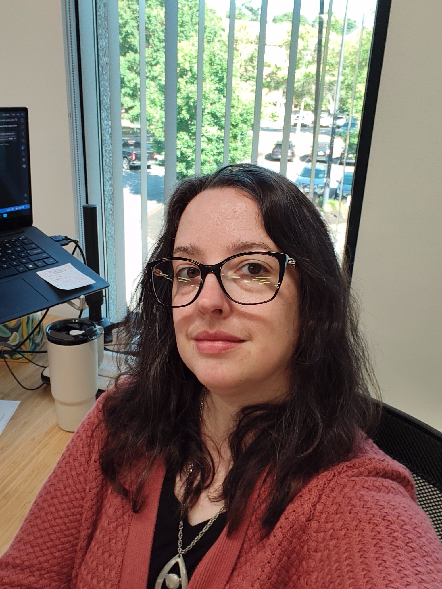 A woman with glasses and long dark hair wearing a pink sweater is sitting at a desk near a window with vertical blinds, with a laptop and a tumbler visible on the desk.