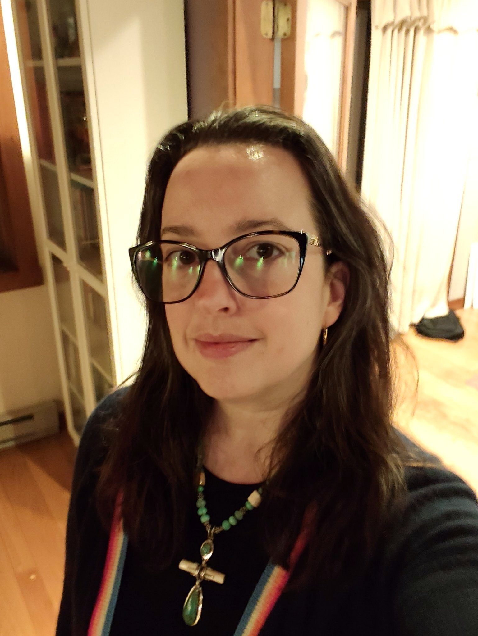 A woman with long dark hair and glasses is wearing a dark top with a colorful striped cardigan and beaded necklaces while standing indoors.