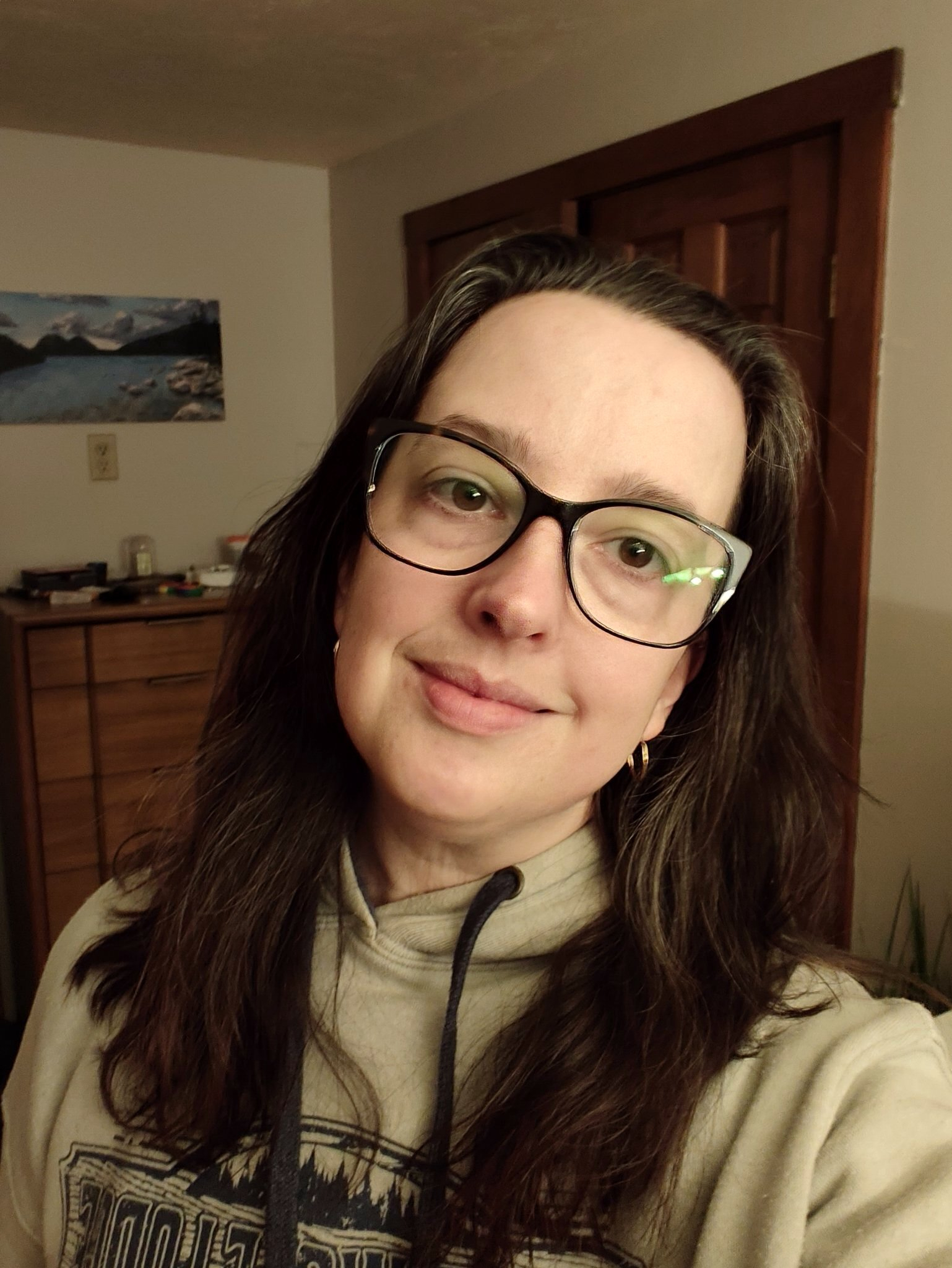 A woman with glasses and long brown hair is smiling softly while wearing a beige hoodie in a warmly lit room.