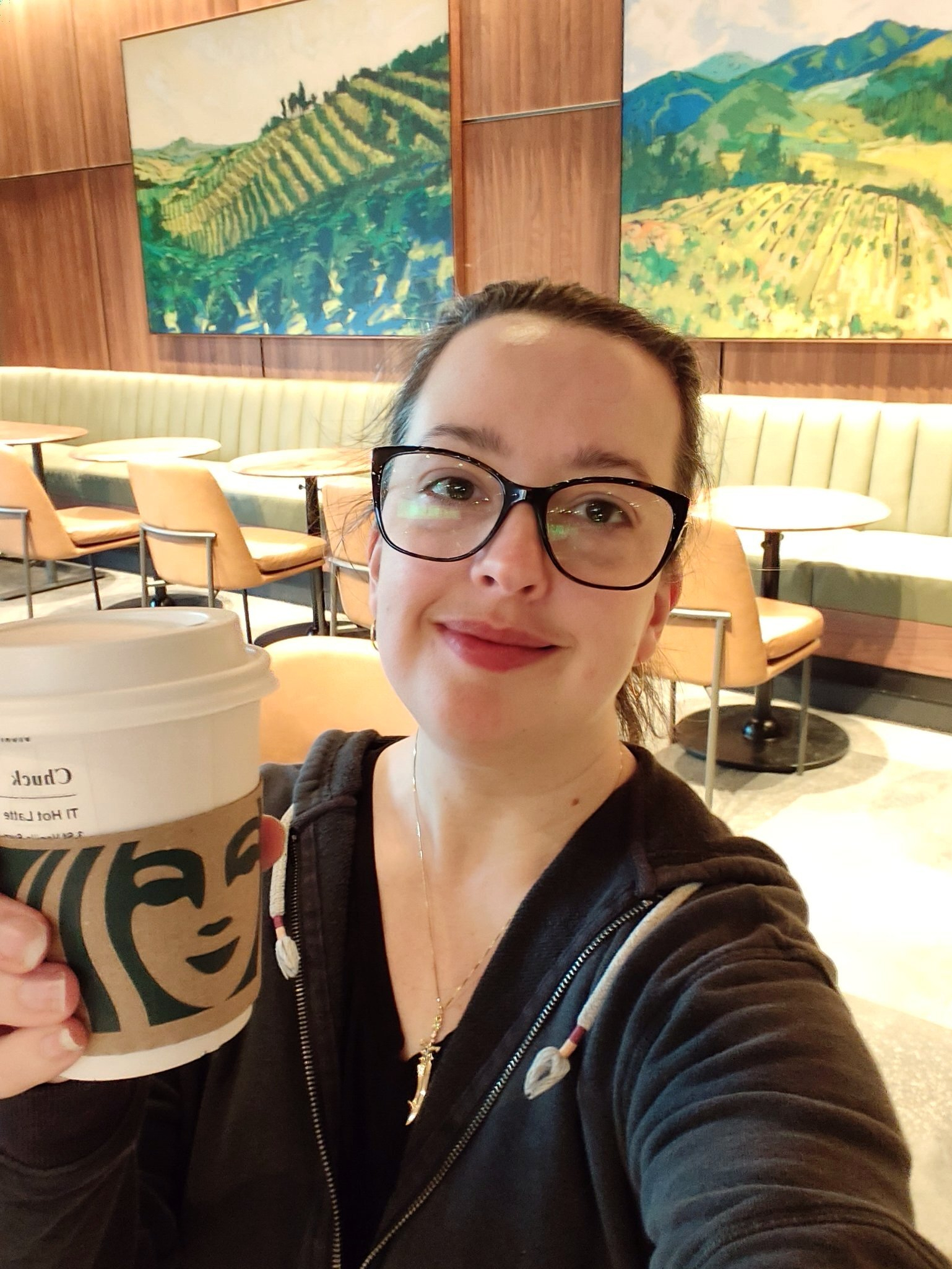 A woman wearing glasses smiles while holding a Starbucks coffee cup inside a café decorated with landscape paintings and modern furniture.