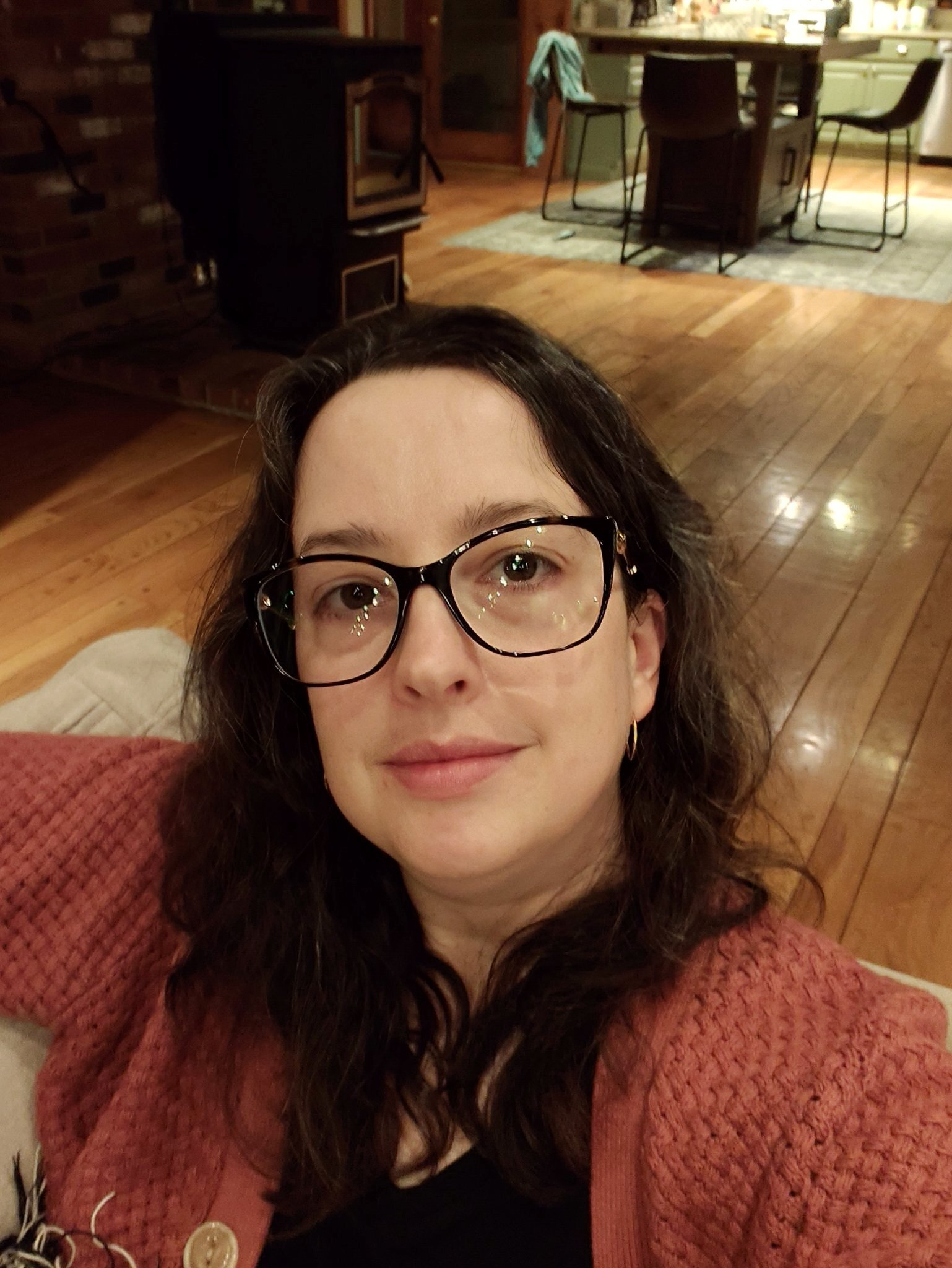 A woman with glasses and dark hair is sitting indoors on a couch, wearing a textured pink cardigan and smiling softly.