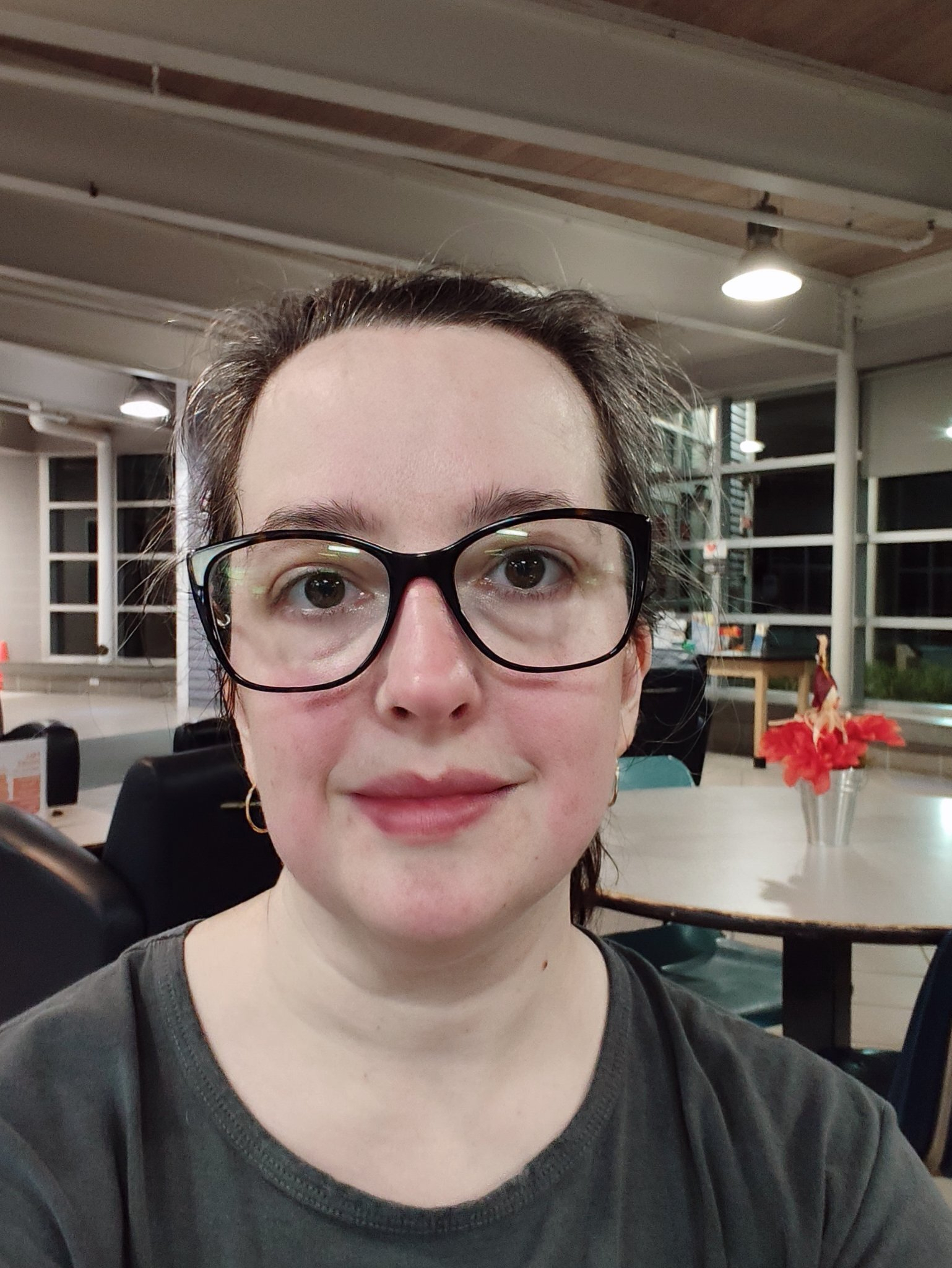 A woman with dark hair and glasses is taking a close-up selfie indoors in a well-lit room with tables, chairs, and a vase with red flowers in the background.