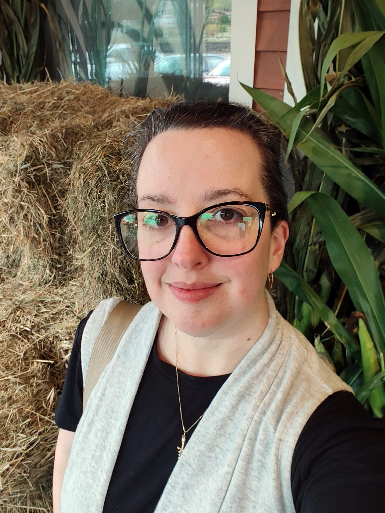 A woman wearing glasses and a light gray vest over a black shirt is smiling in front of some hay and green plants.