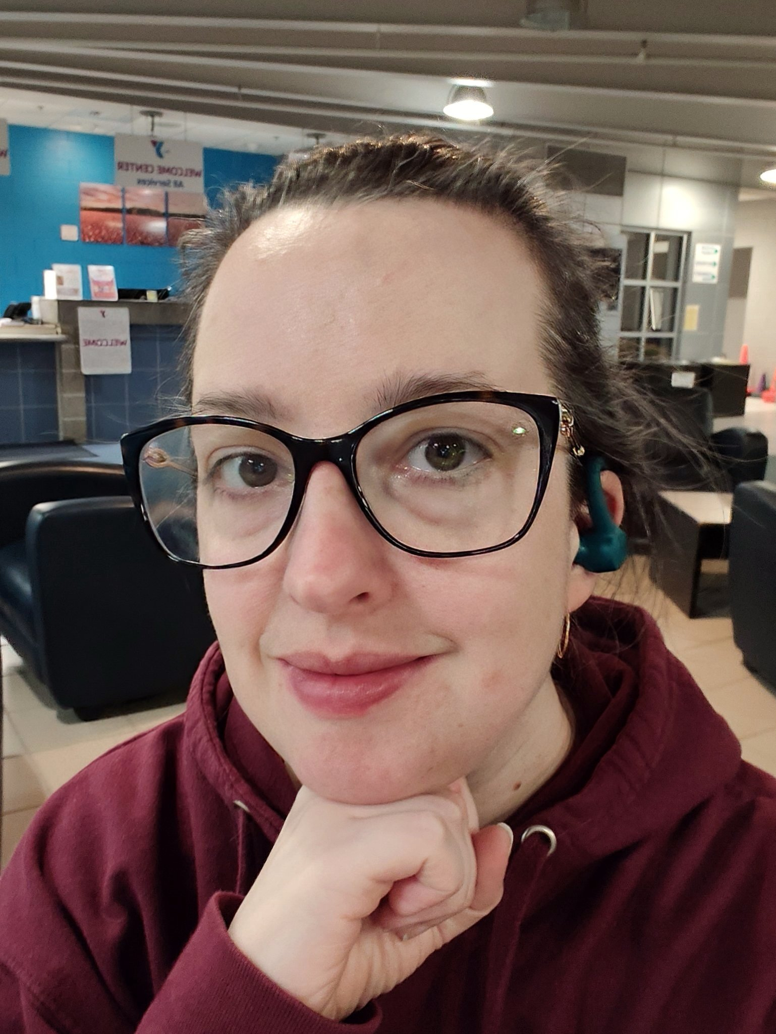 A person wearing large black glasses and a maroon hoodie is resting their chin on their hand while sitting in a lounge area.