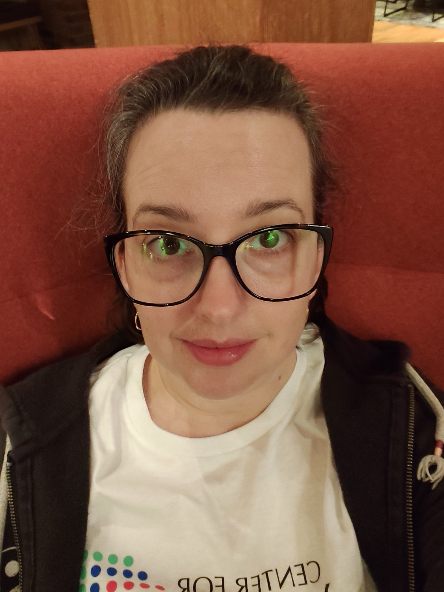 A person with glasses and dark hair is sitting against a red cushioned background, wearing a white shirt and a dark jacket.