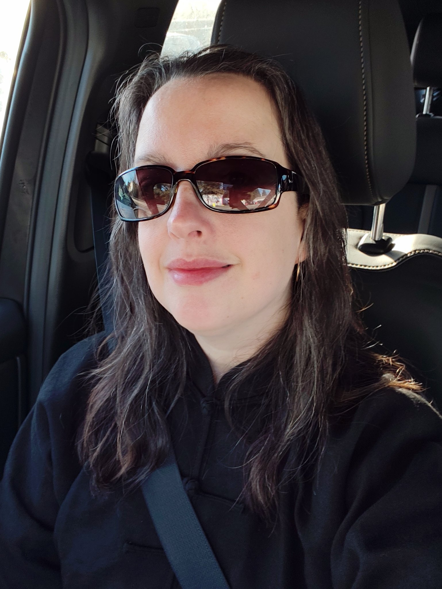 A woman wearing dark sunglasses and a black top is sitting inside a car with her seatbelt fastened.
