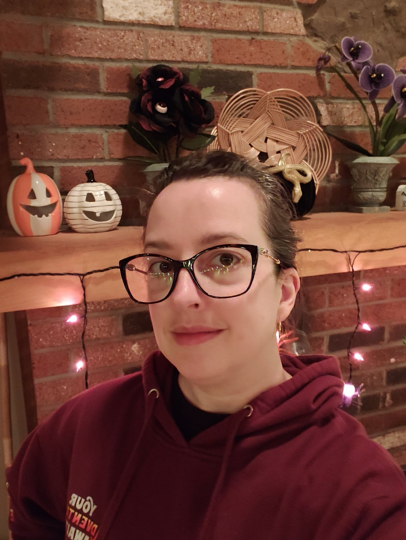 A person wearing glasses and a maroon hoodie is taking a selfie in front of a brick wall decorated with Halloween-themed items and string lights.
