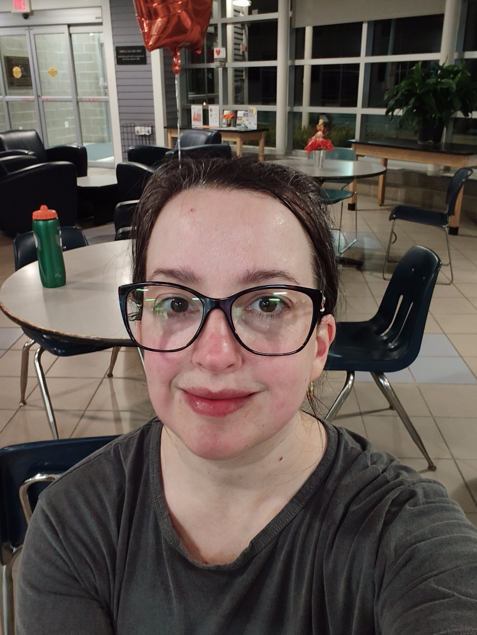 A person wearing glasses and a dark shirt is taking a selfie in a room with tables, chairs, and a green water bottle in the background.