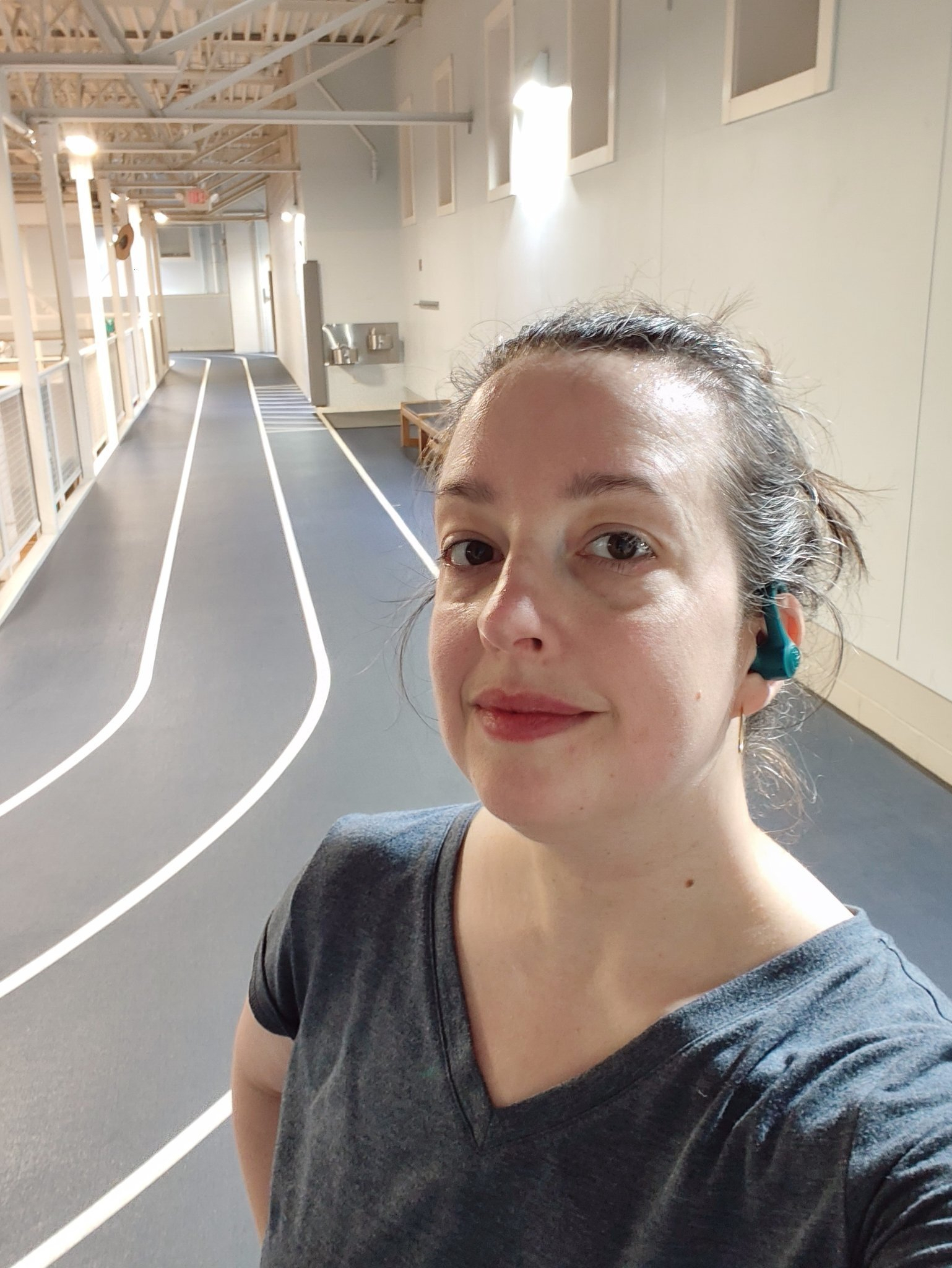 A woman wearing a gray V-neck shirt and blue earbuds is taking a selfie on an indoor running track with lane markings.