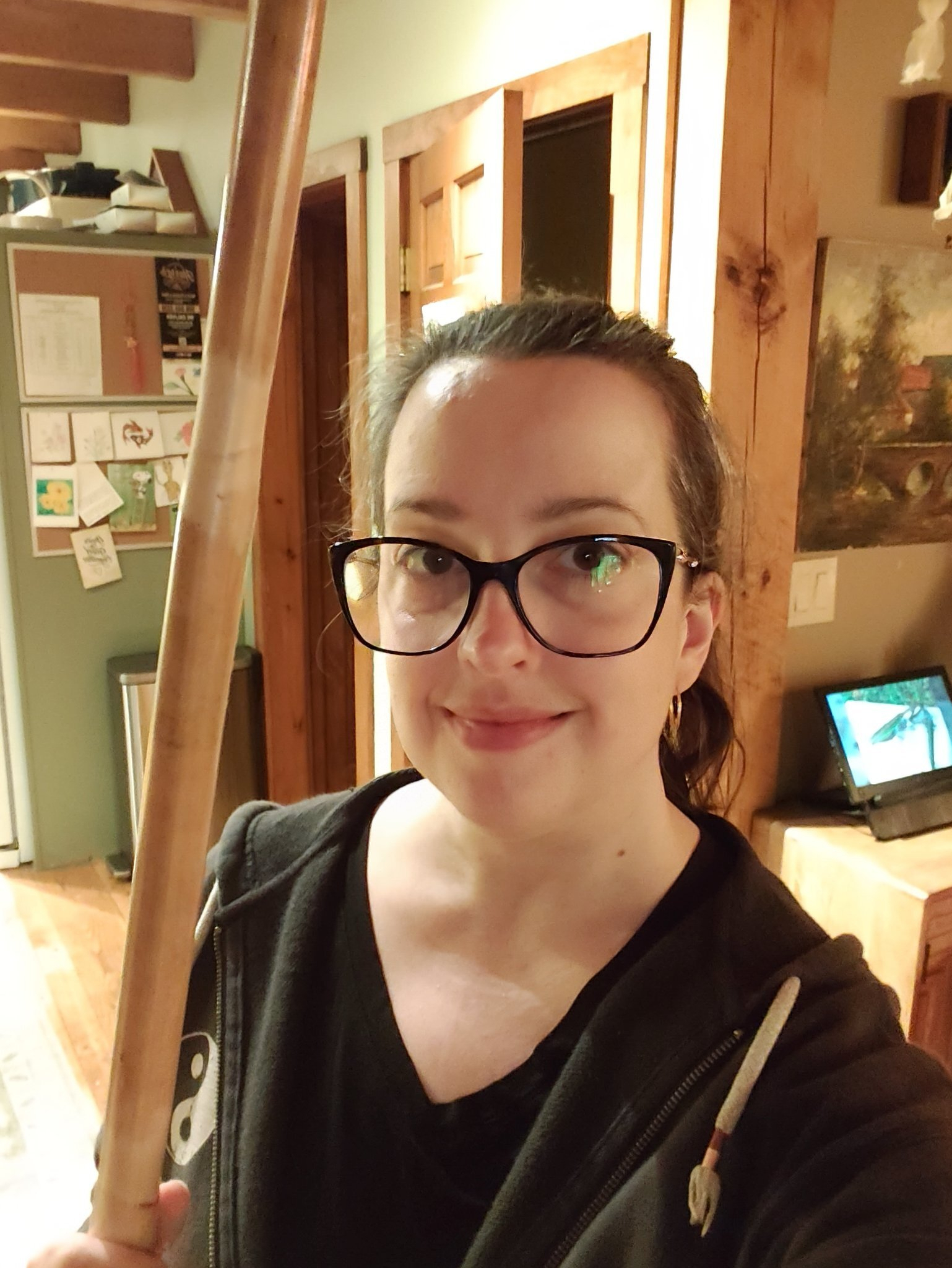 A woman wearing glasses and a black hoodie is smiling while holding a long wooden stick inside a cozy room with wooden accents.