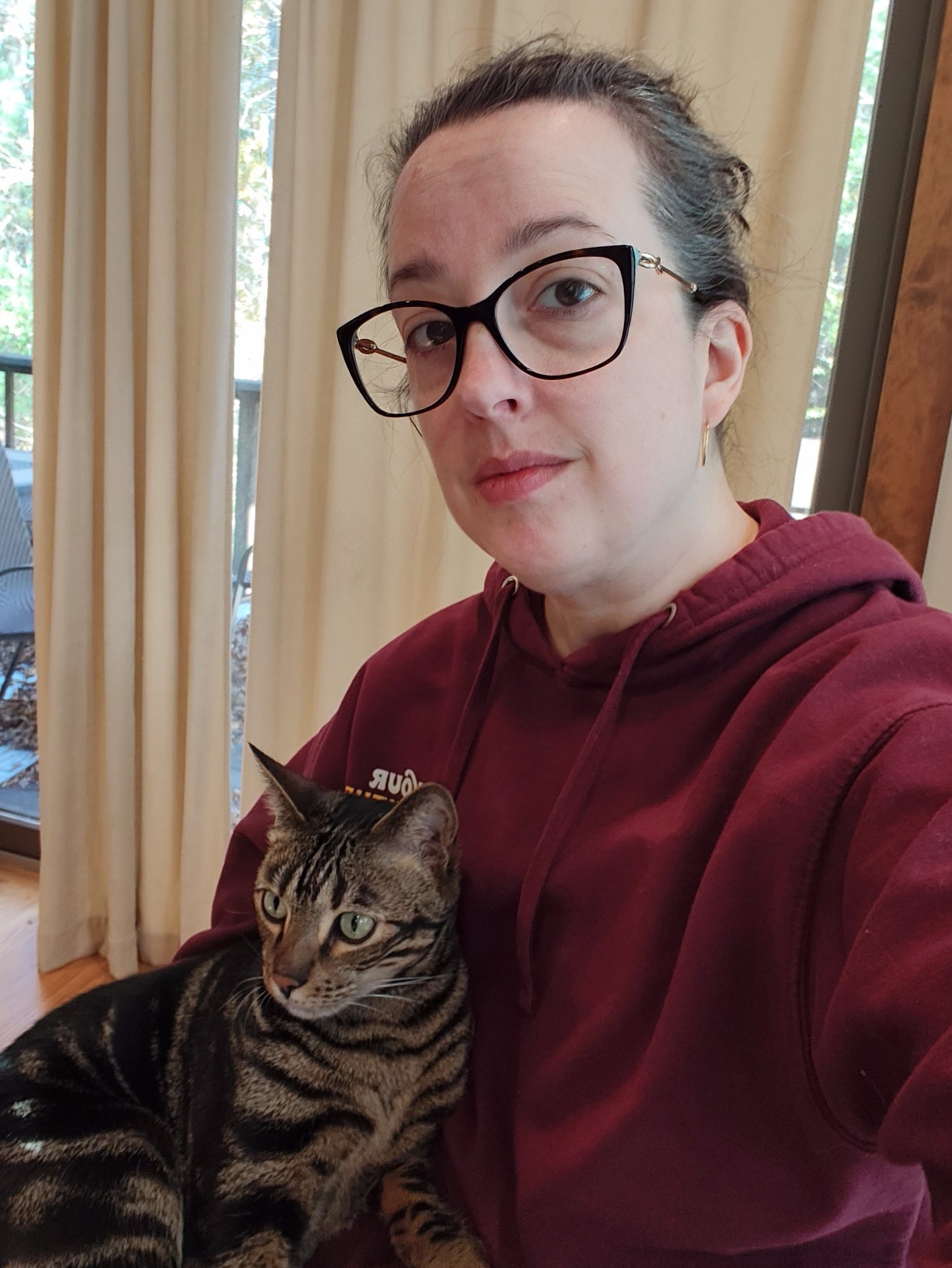 A person wearing glasses and a maroon hoodie is sitting indoors with a tabby cat resting on their lap.