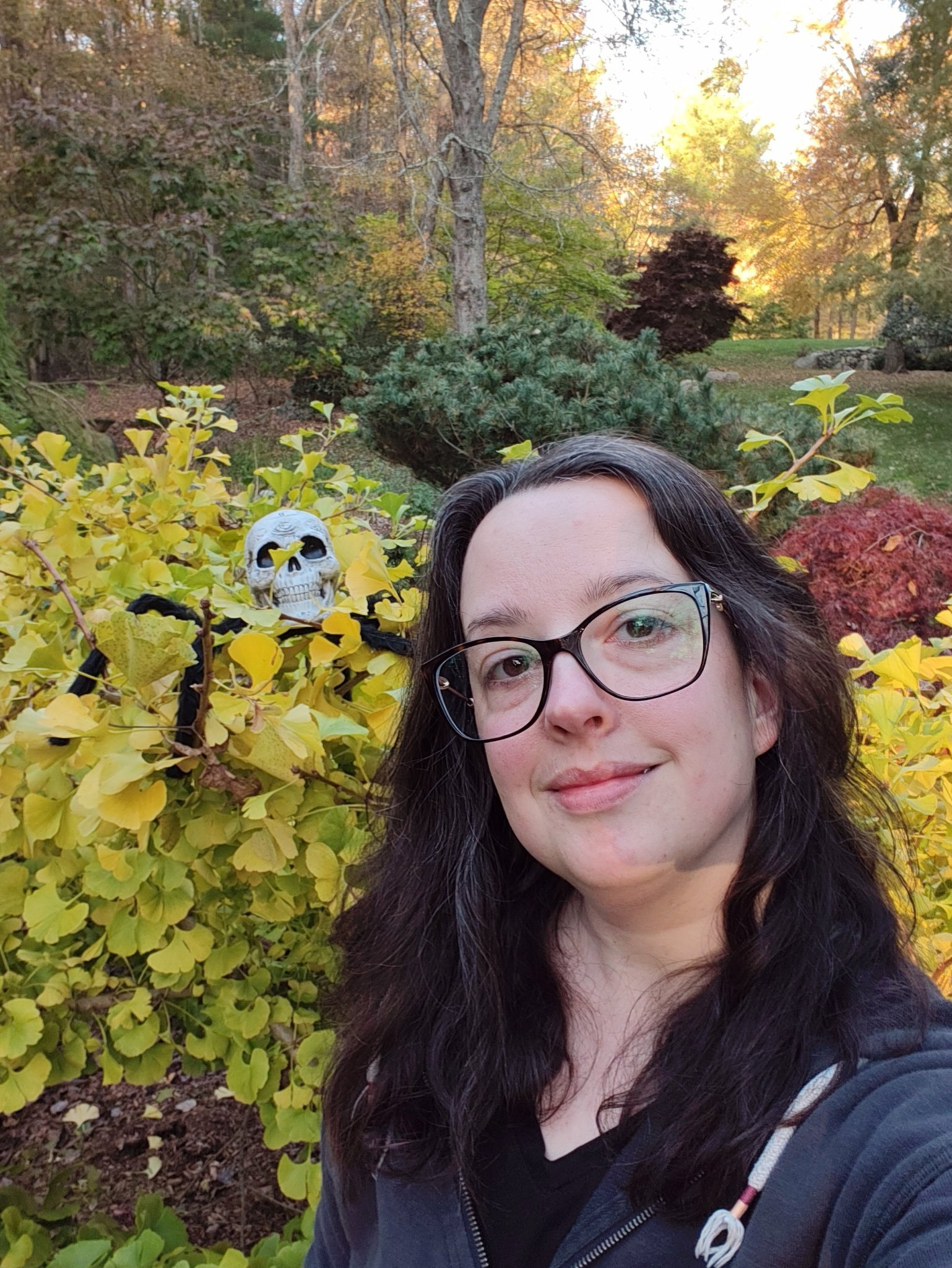 A woman with glasses takes a selfie outdoors near bushes, one of which is decorated with a Halloween skull and spider.