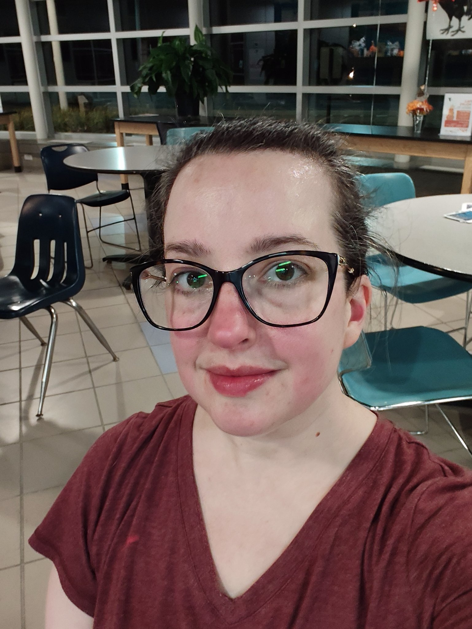 A person with glasses and a red shirt is smiling indoors, with tables and chairs in the background.