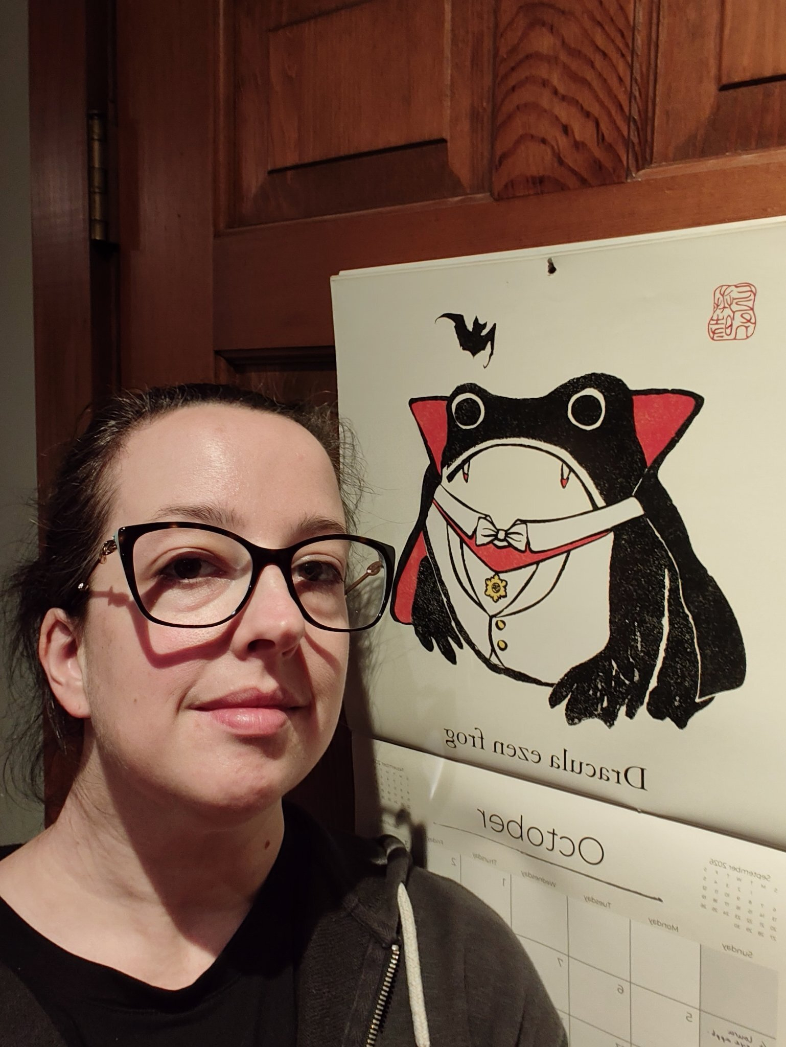 A person wearing glasses is standing next to a calendar featuring a cartoon frog dressed as a vampire with the caption Dracula eezan frog.