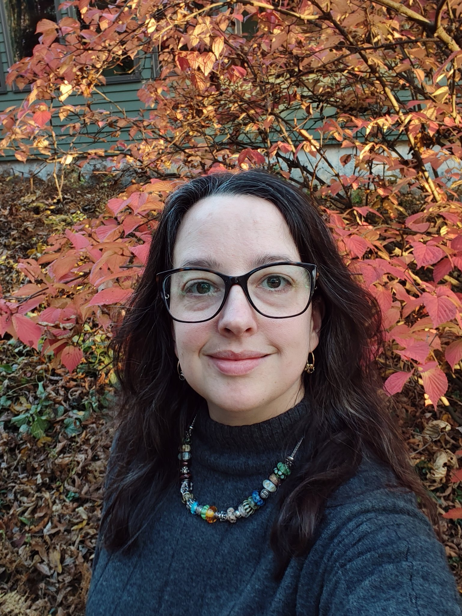 A woman with glasses and long dark hair is smiling while standing outdoors in front of colorful autumn foliage.