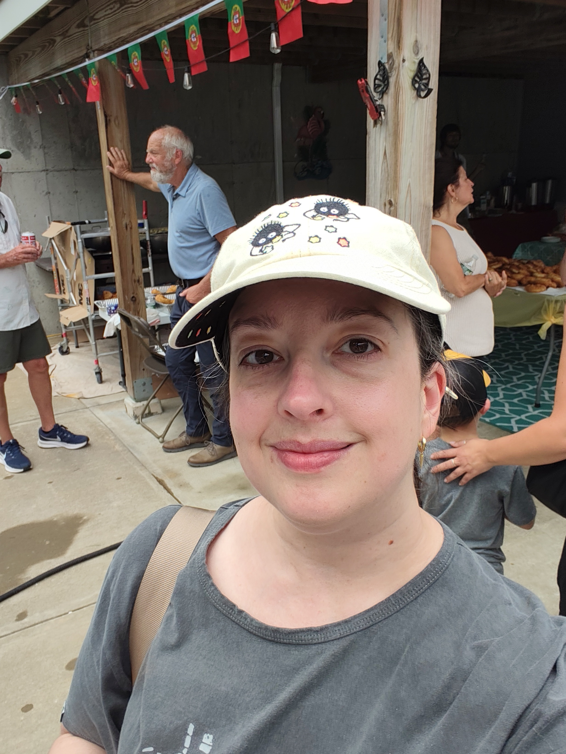 A woman wearing a cap with cartoon eyes takes a selfie at an outdoor gathering where people are socializing and food is served under a decorated wooden canopy.