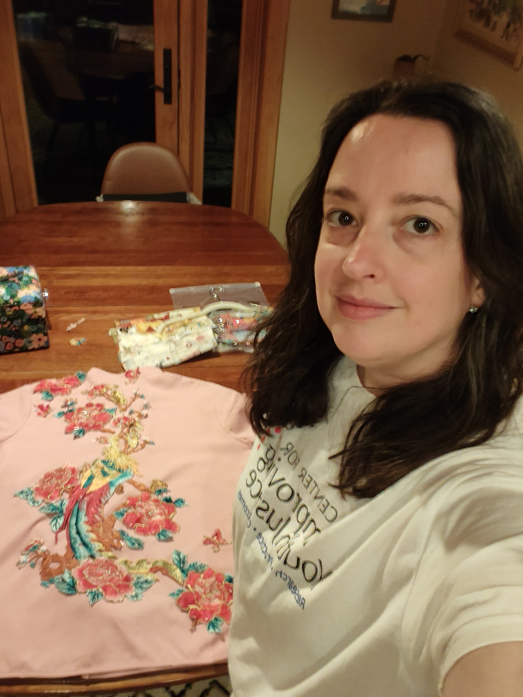 A woman with long dark hair is taking a selfie indoors, standing next to a wooden table with a pink garment featuring colorful floral embroidery laid out on it.