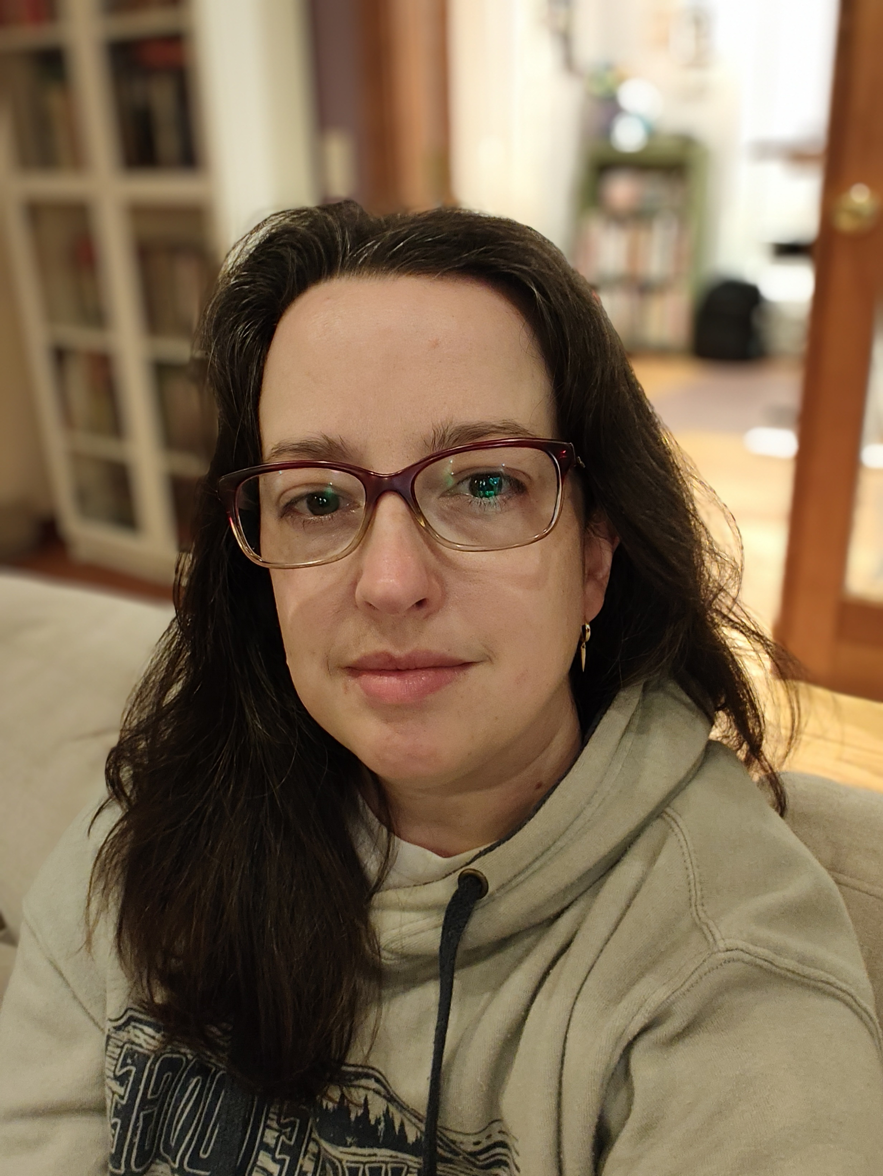 A woman with long dark hair, wearing glasses and a beige hoodie, is sitting indoors with bookshelves and a door in the background.