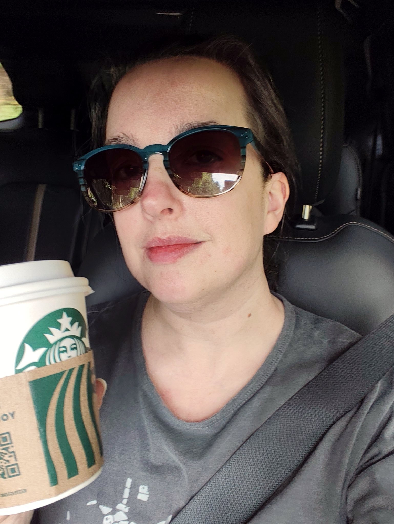 A woman wearing sunglasses and a seatbelt is holding a Starbucks coffee cup while sitting inside a car.