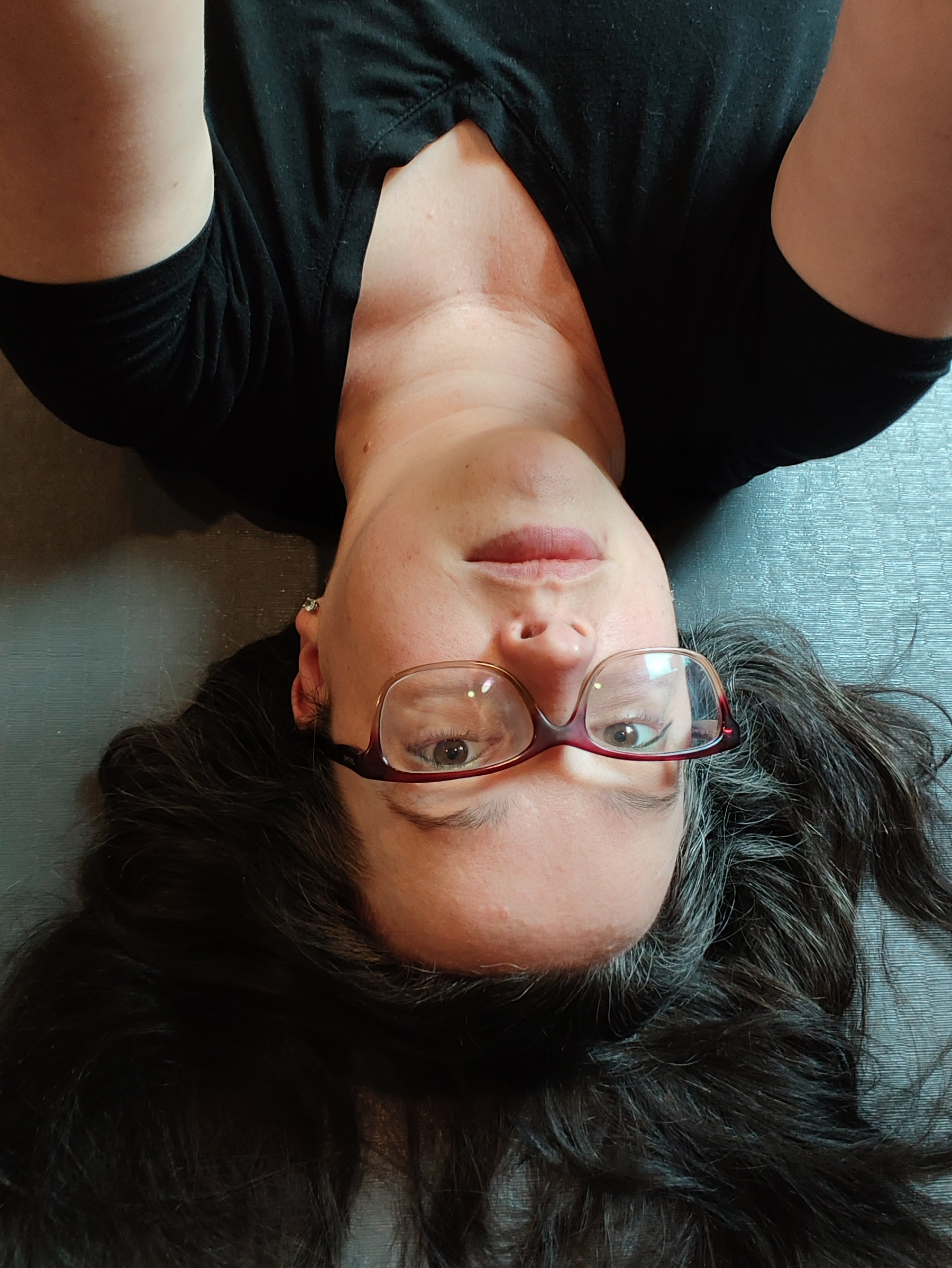 A person with glasses and long dark hair is lying down on a textured surface, wearing a black top.