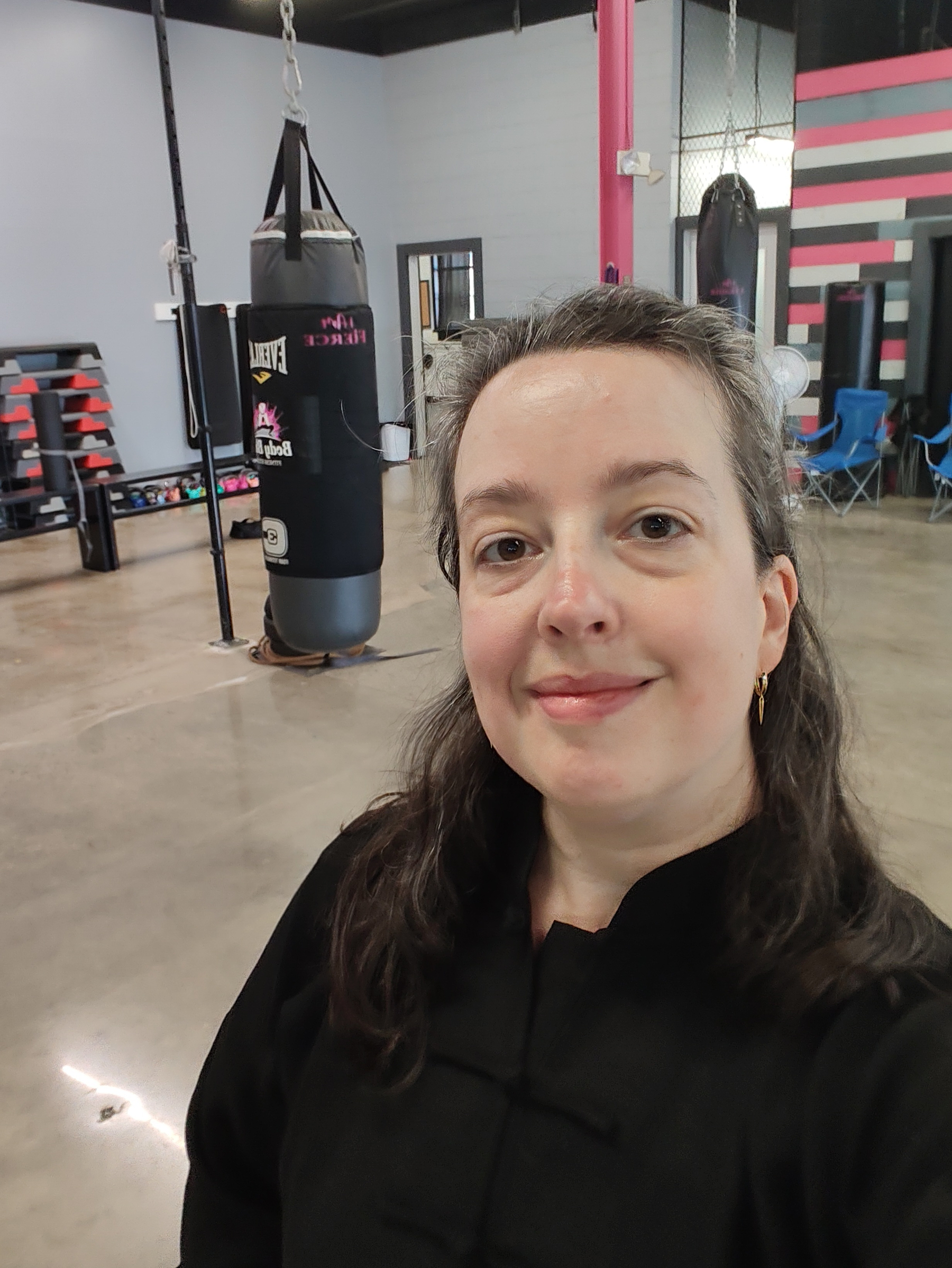 A woman in a martial arts uniform is taking a selfie inside a gym with punching bags and exercise equipment in the background.