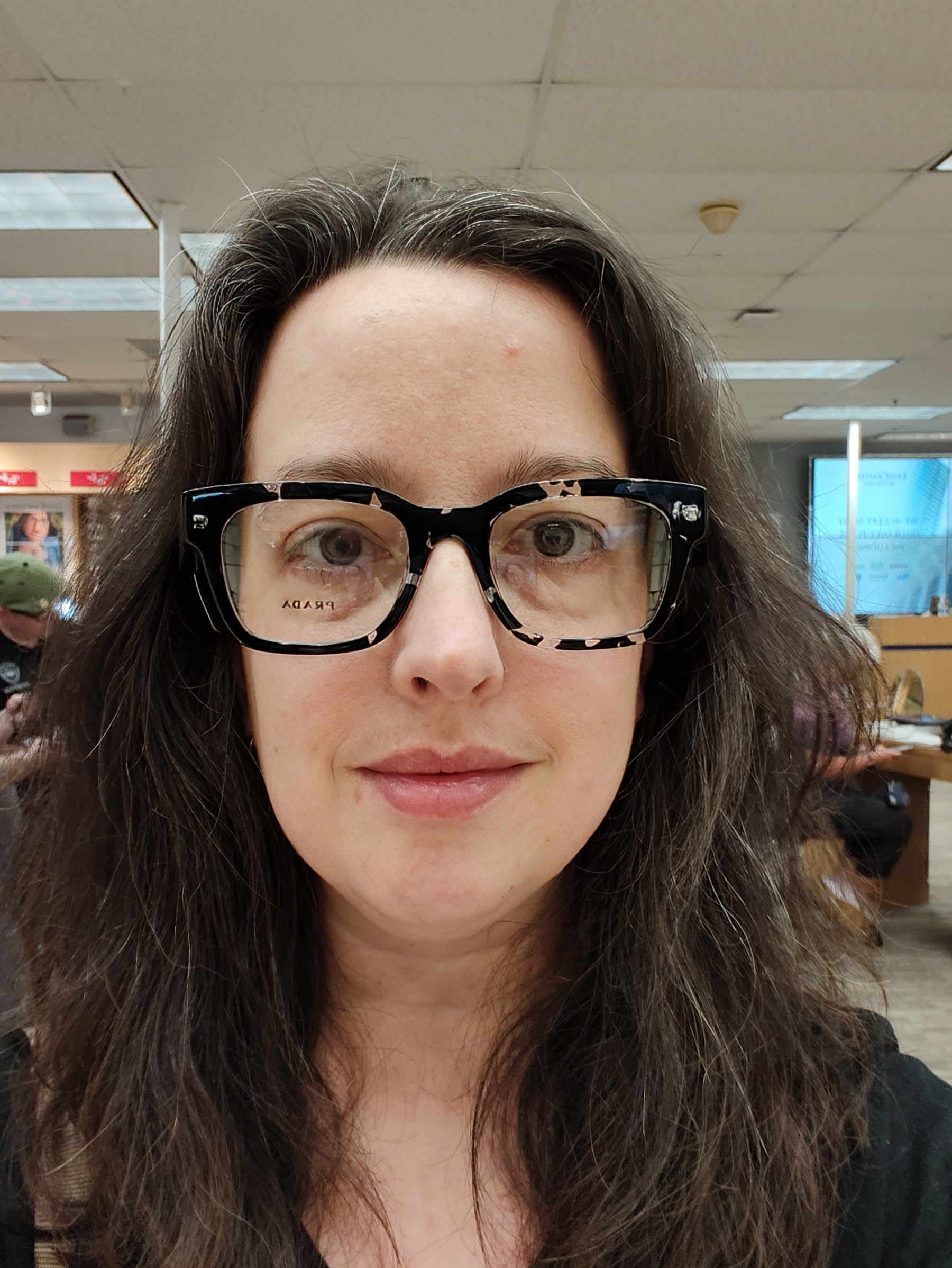 A woman with long, wavy hair is wearing large, black-framed glasses and is looking directly at the camera in an indoor setting.