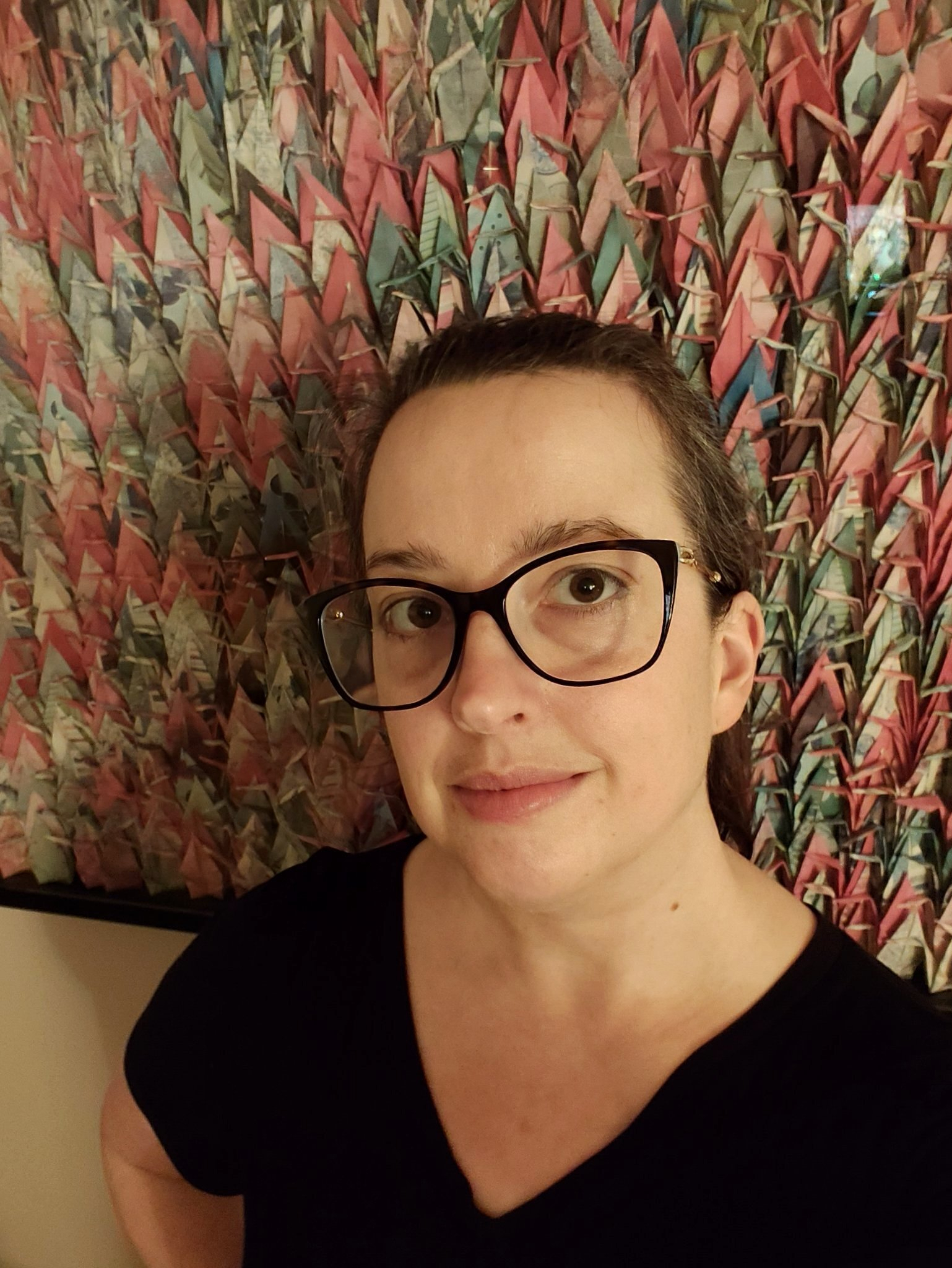 A woman wearing glasses and a black shirt stands in front of an intricate and colorful origami crane collage.