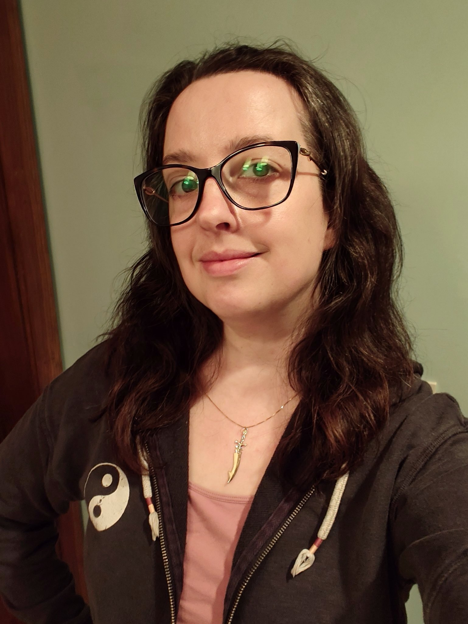 A person with long dark hair, wearing glasses, a necklace, and a zip-up hoodie with a yin-yang symbol is smiling slightly at the camera.