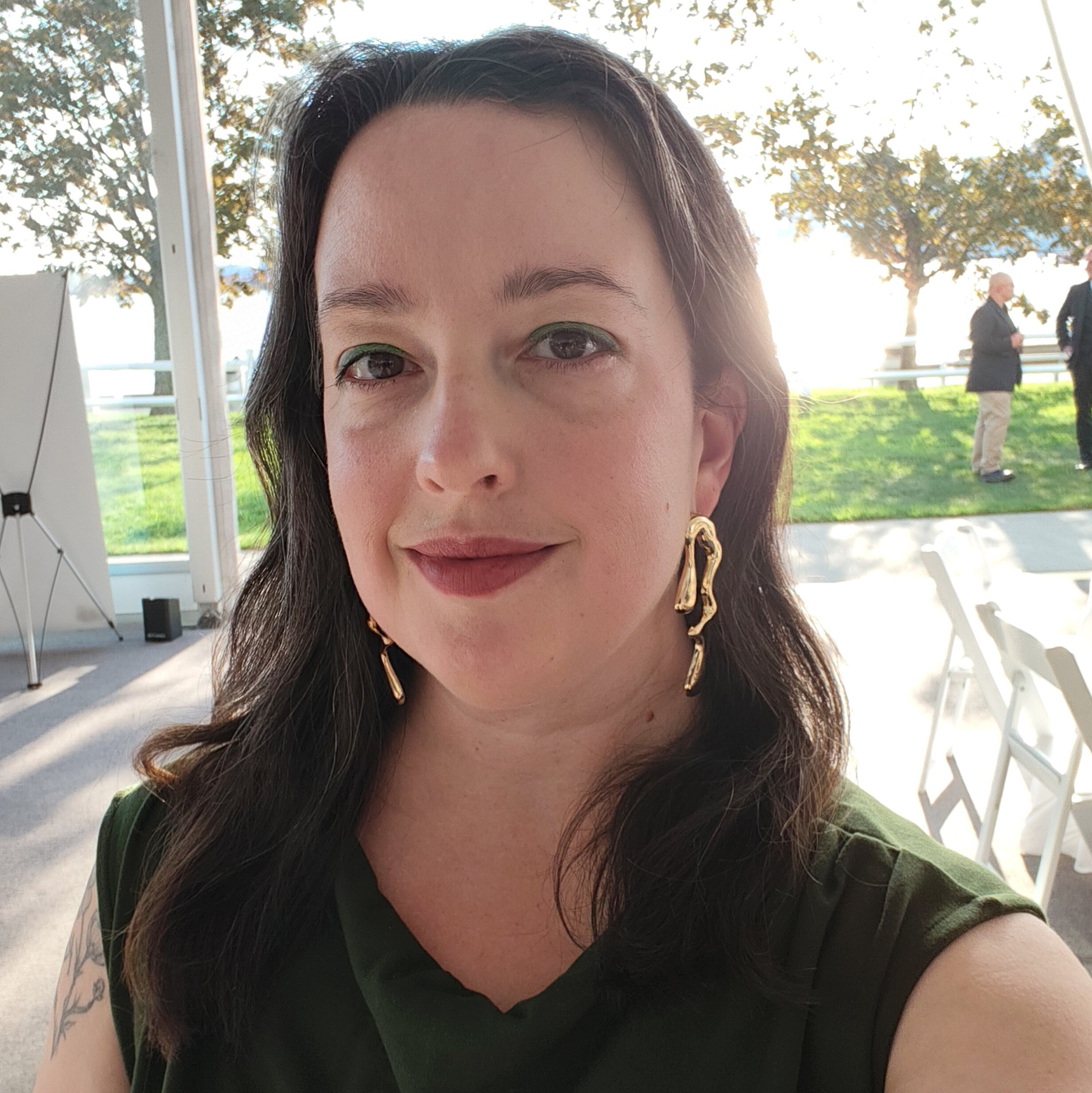 A woman with long dark hair, wearing a green top and unique gold earrings, takes a selfie in a well-lit outdoor space with white chairs and a grassy background.