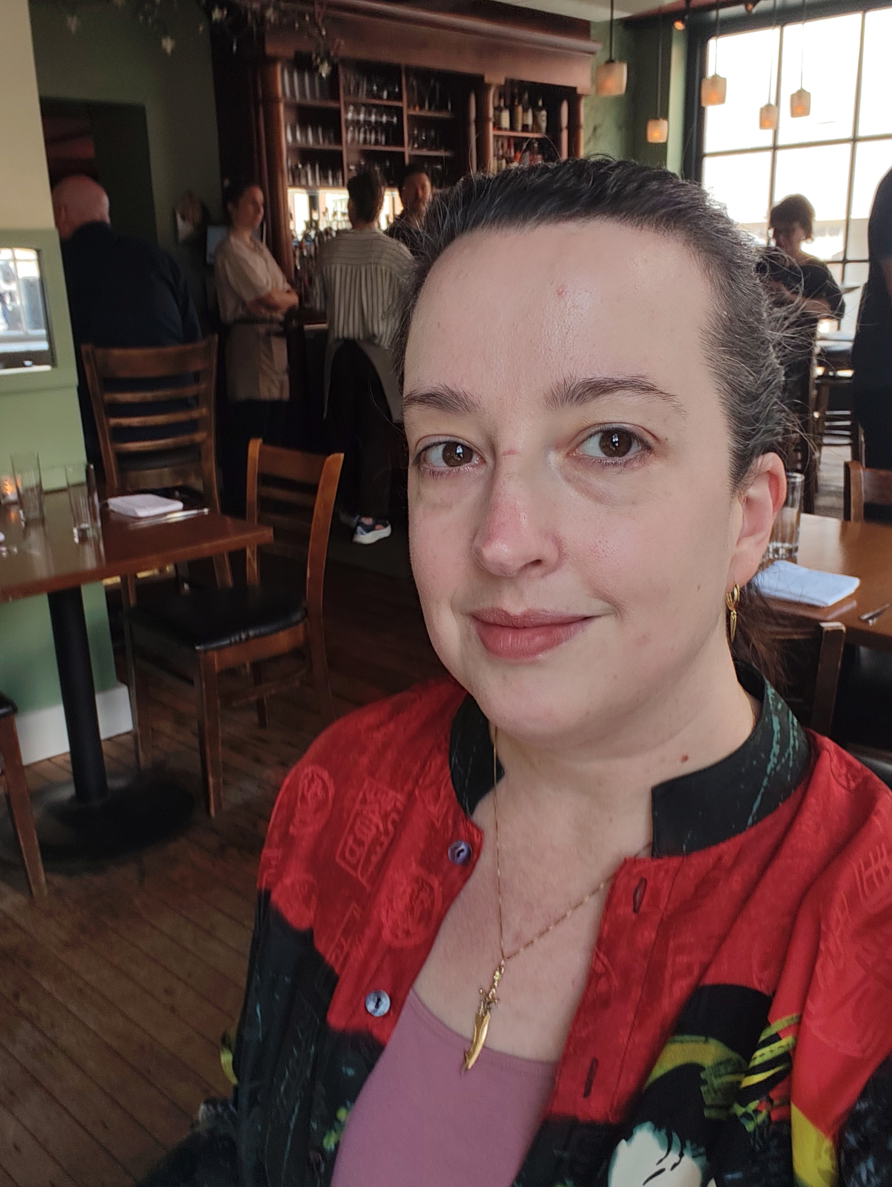 A person with a red and black patterned shirt is smiling while taking a selfie inside a warmly lit restaurant with wooden floors and other people in the background.