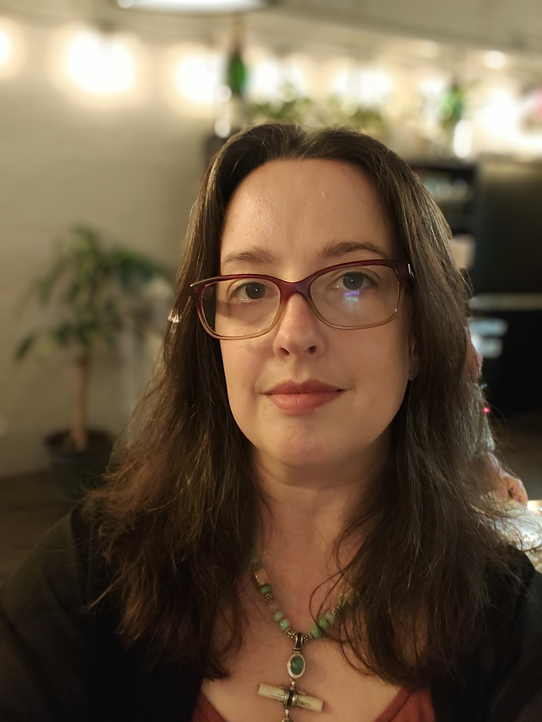 A woman with long brown hair and glasses is wearing a necklace with a cross pendant, sitting indoors with a softly blurred background featuring plants and warm lighting.