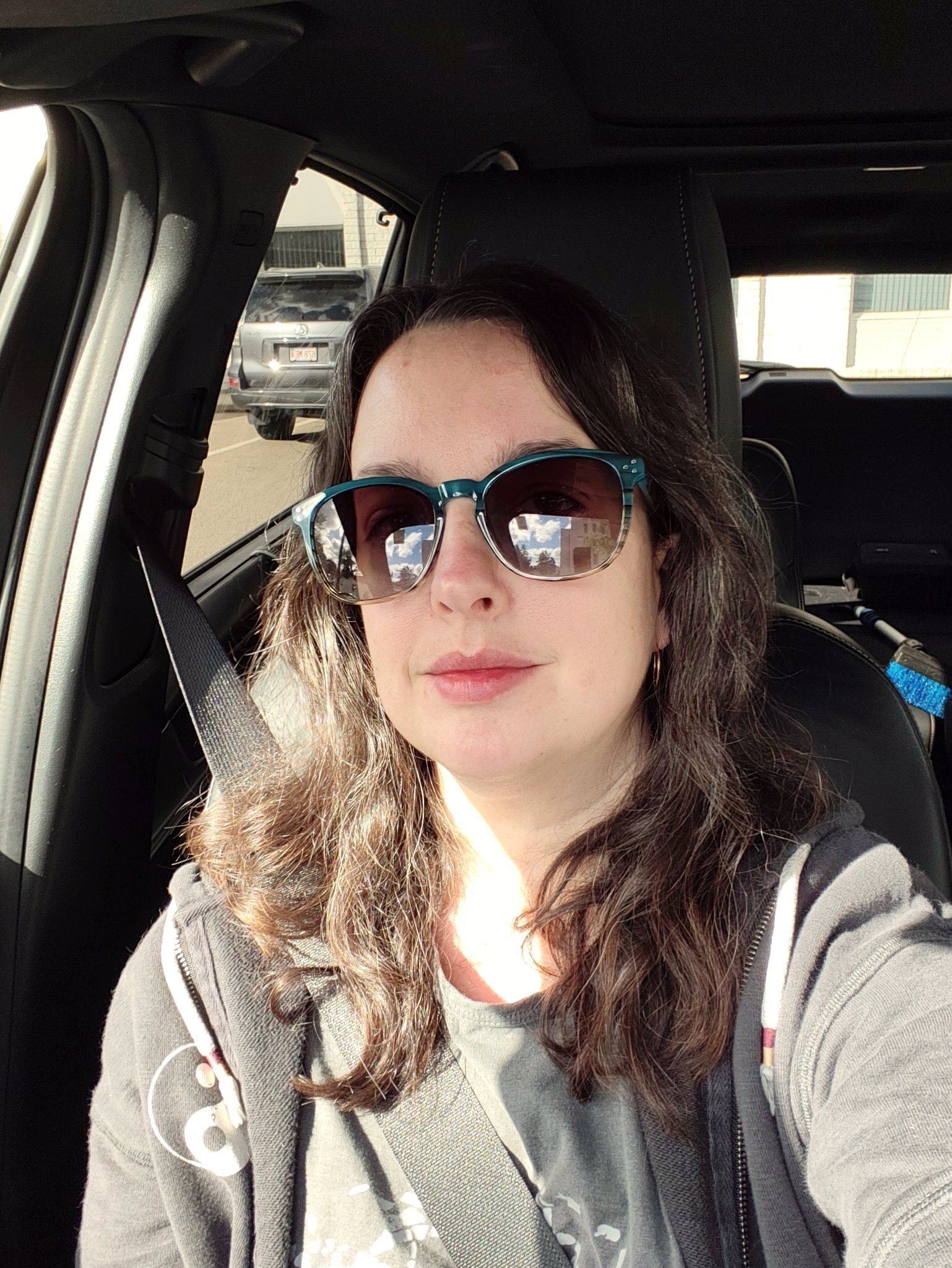 A woman wearing teal sunglasses and a dark hoodie is sitting in the driver's seat of a car with sunlight illuminating part of her face.