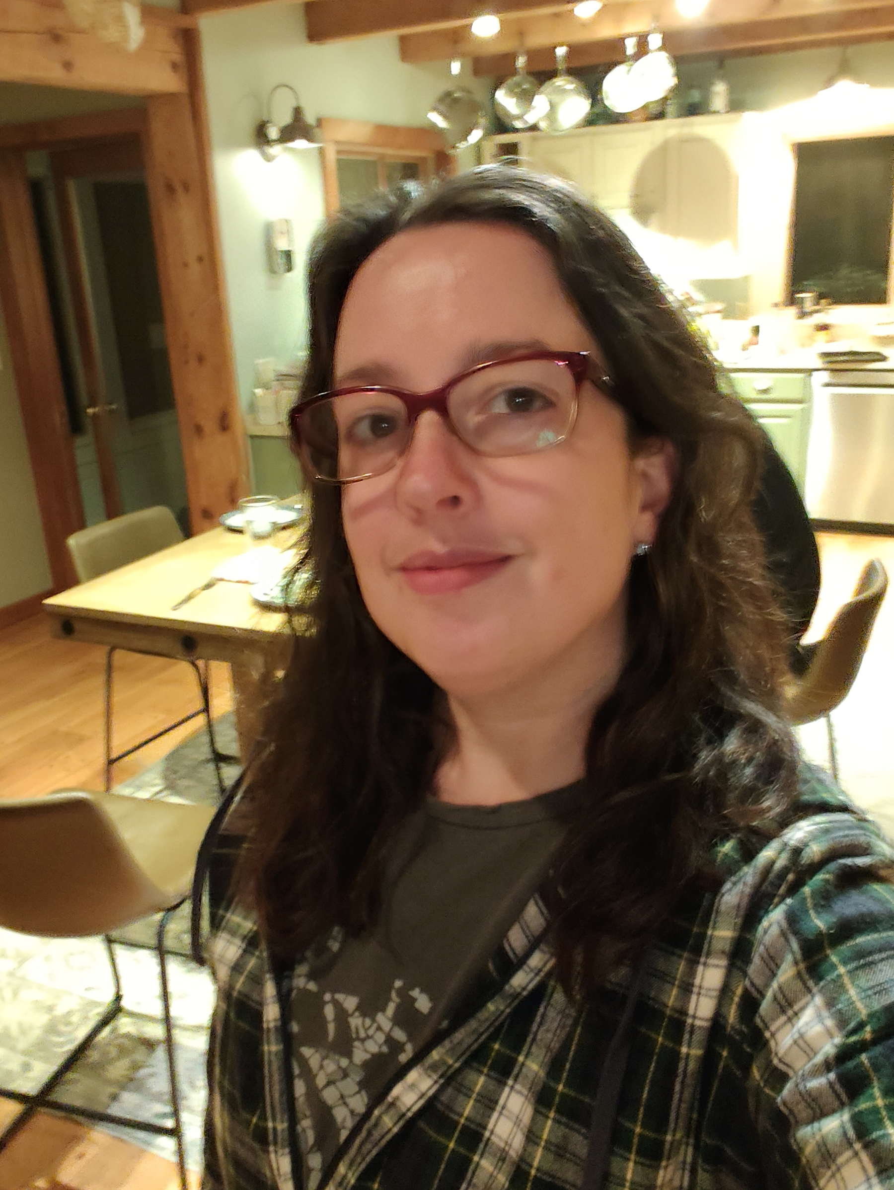 A woman with glasses and long dark hair is smiling and taking a selfie in a warmly lit kitchen and dining area.
