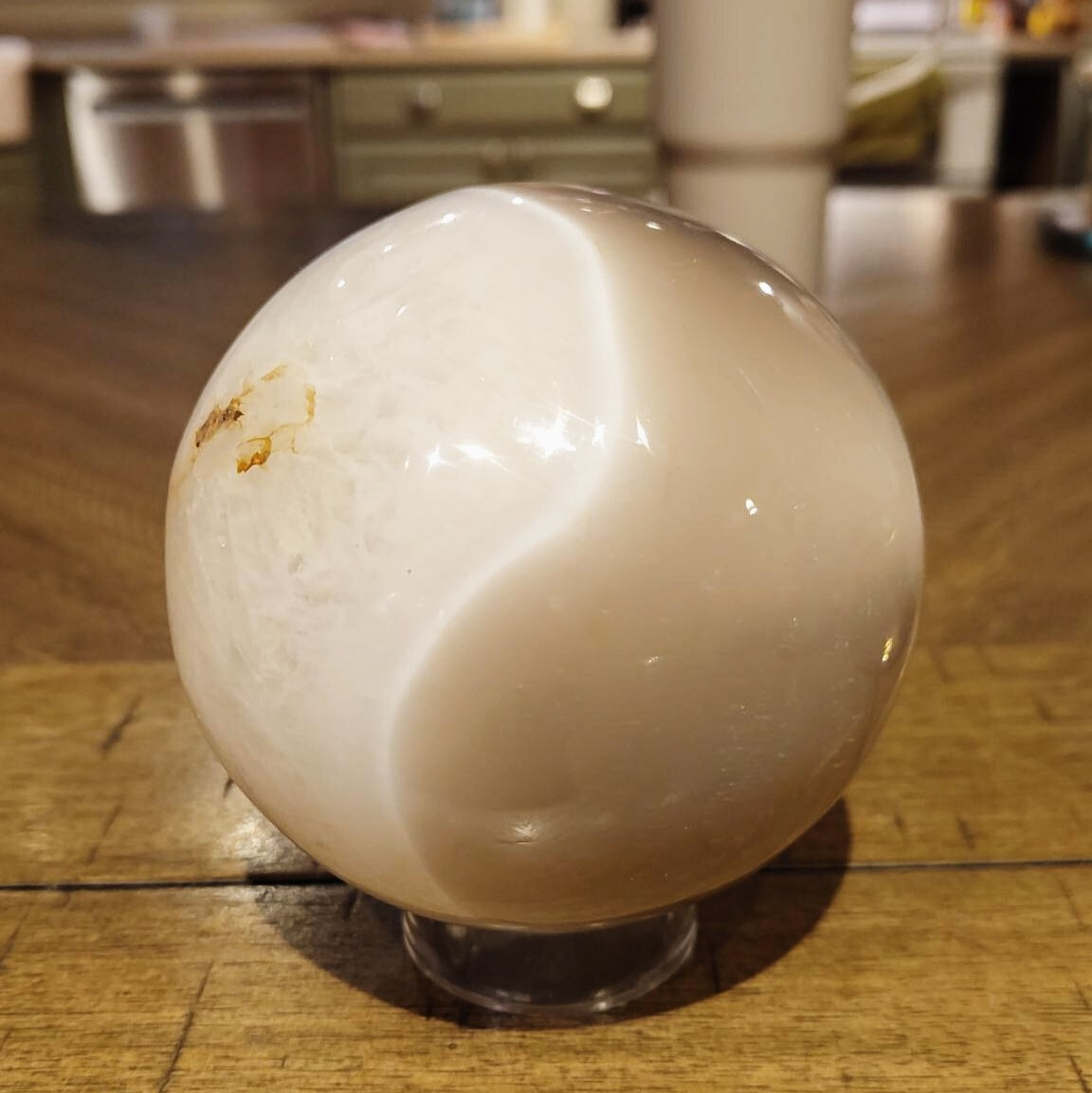 A smooth, polished white stone sphere with a subtle beige pattern rests on a clear stand on a wooden surface.