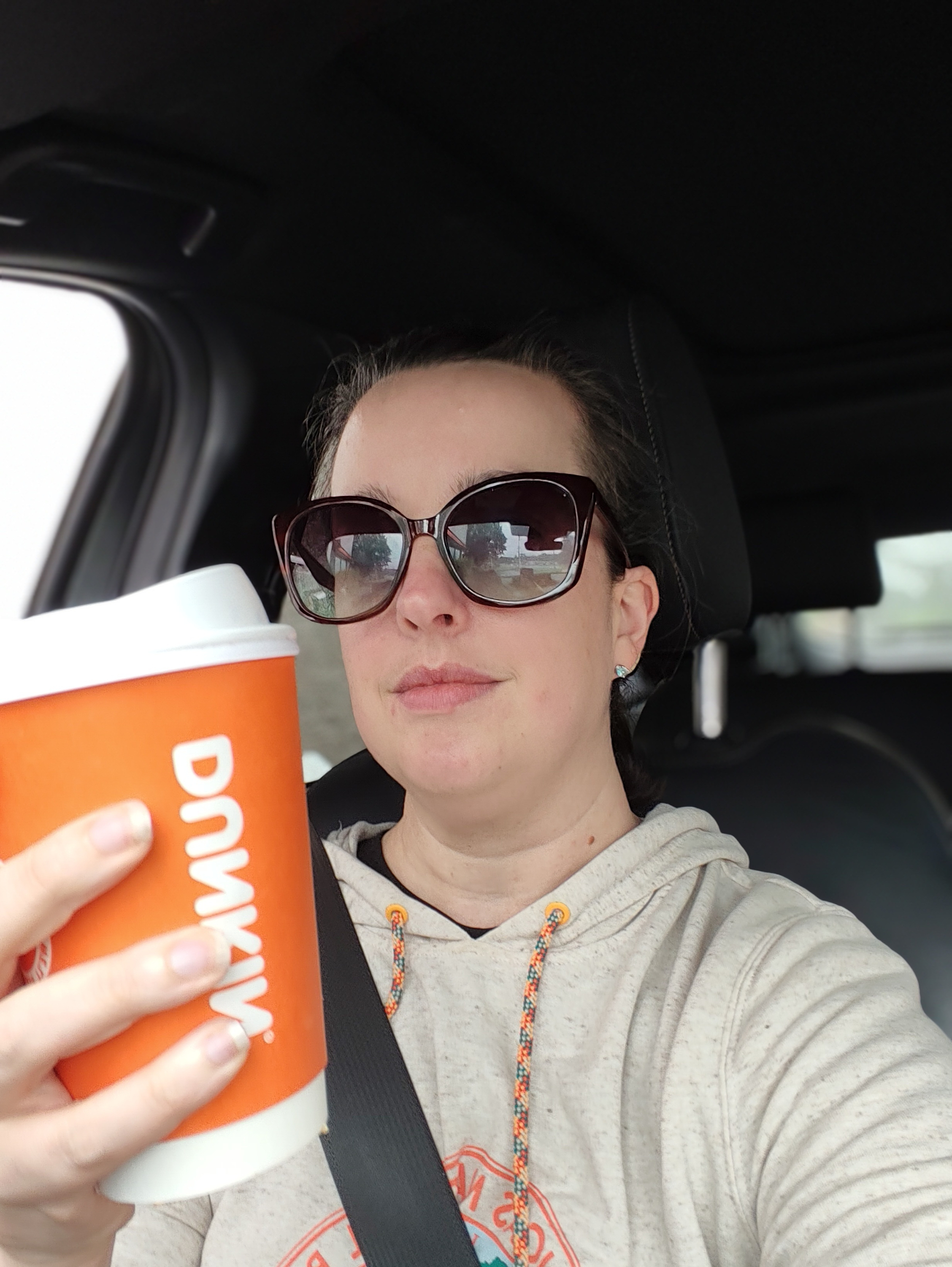A woman wearing large sunglasses and a light hoodie is holding a Dunkin' coffee cup while seated in a car.