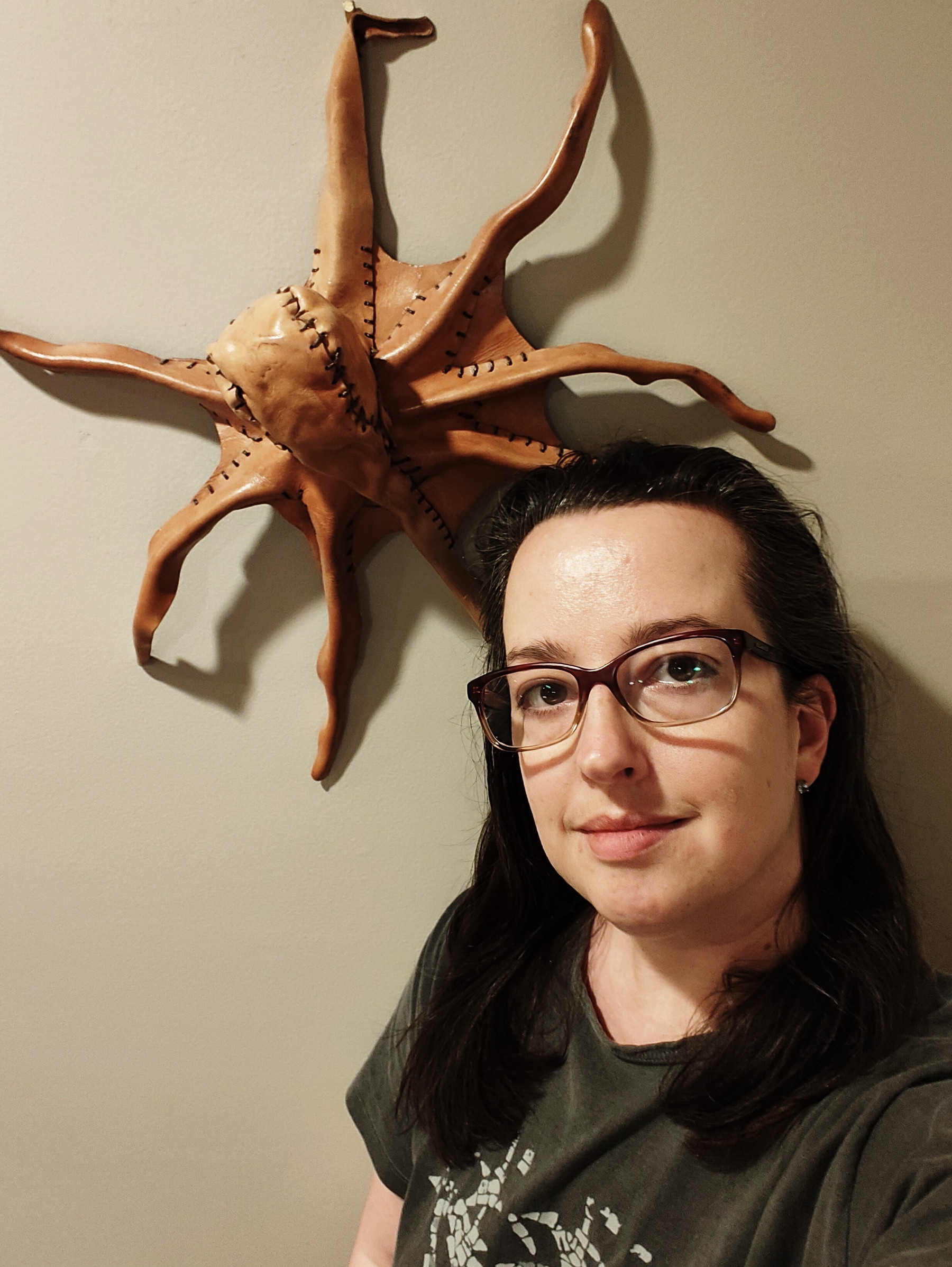 A person wearing glasses is posing in front of a wall-mounted sculpture resembling an octopus made from stitched leather or similar material.