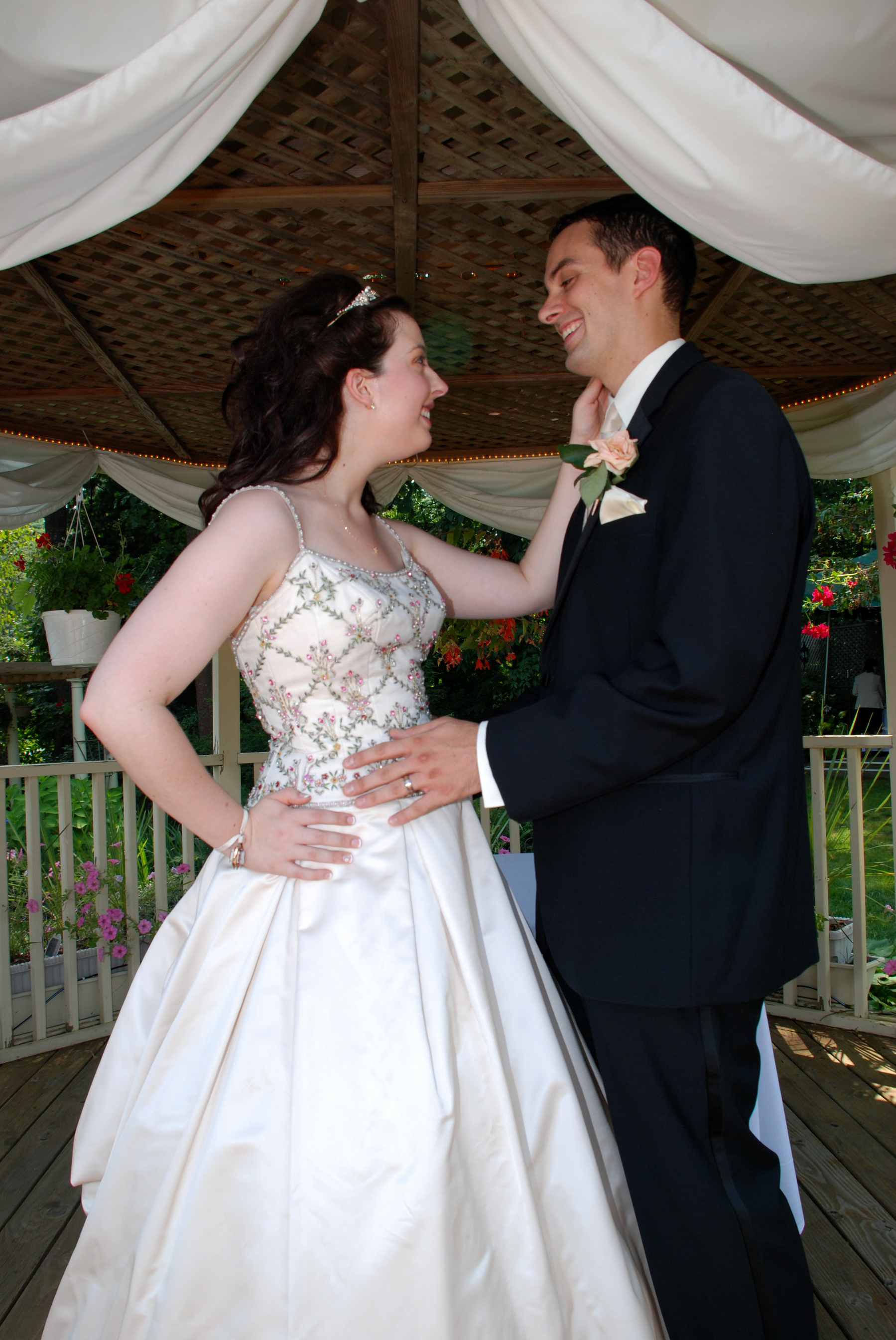 A bride and groom are smiling and affectionately looking at each other while standing under a decorated gazebo.