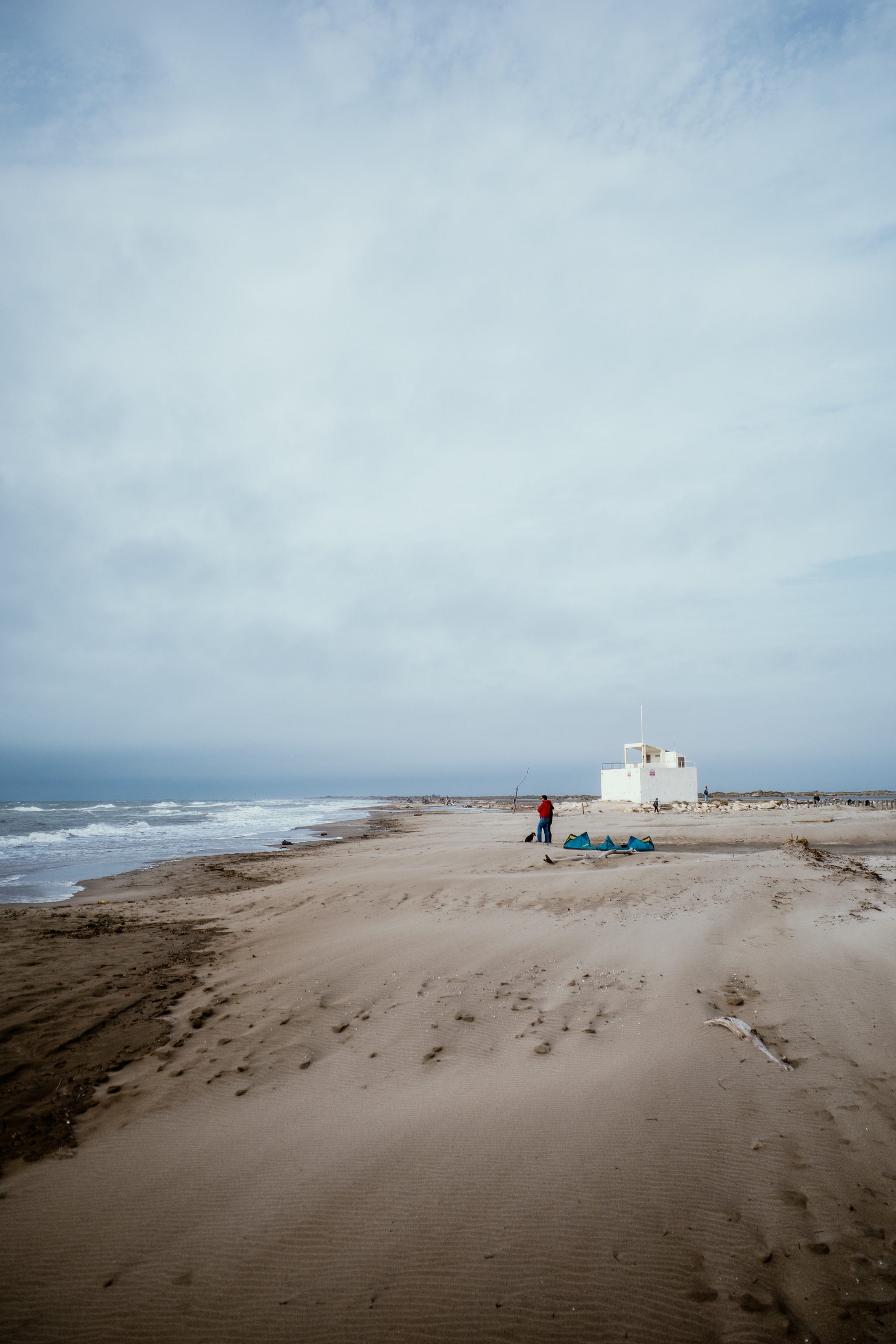 The image shows a solitary person with a dog standing on a wide, deserted beach, with a white building in the distance and waves gently crashing on the shore under a cloudy sky.