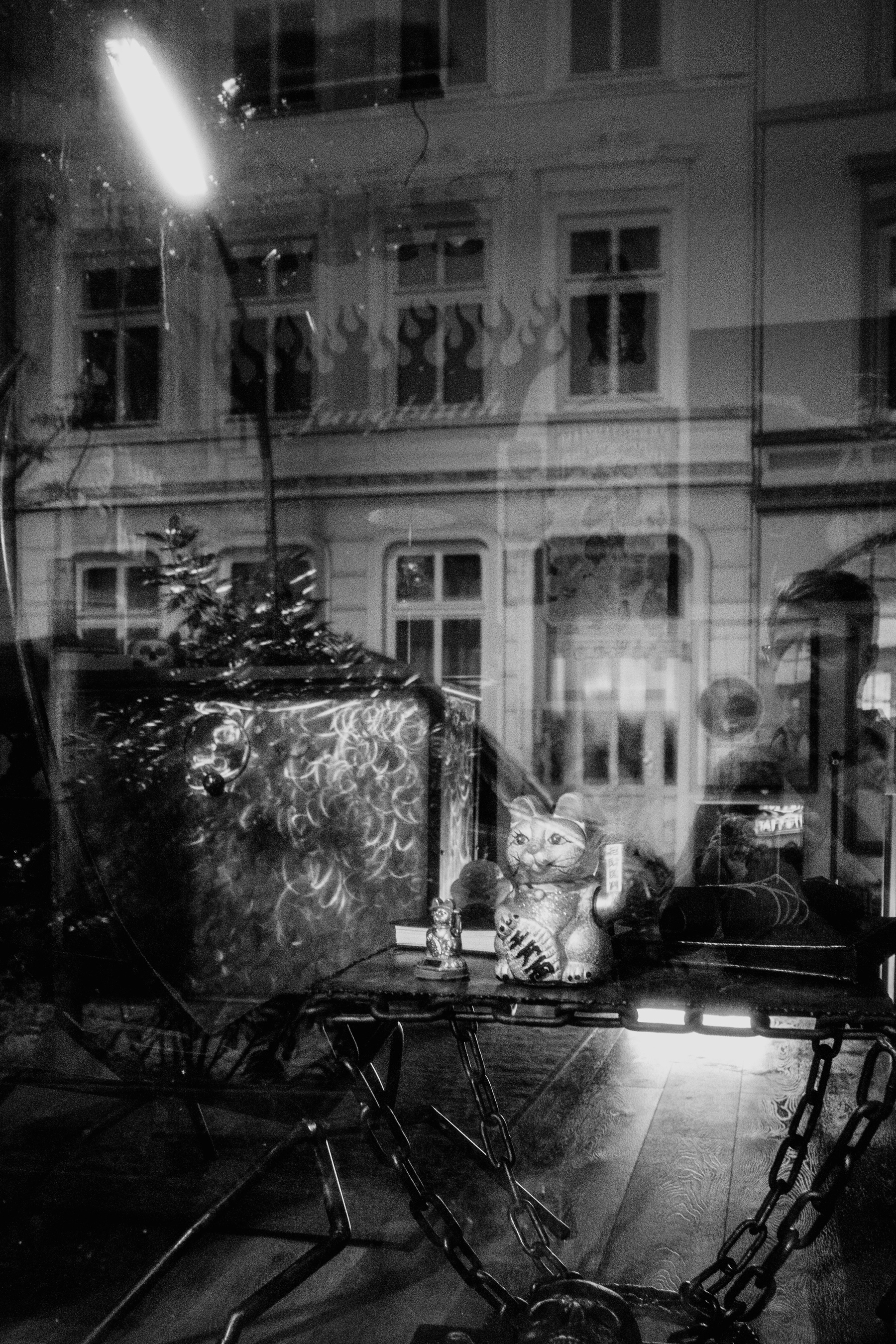 A black-and-white photo capturing a storefront window display that includes a maneki-neko (beckoning cat) statue and other objects, with reflections of a building facade and a person visible in the glass.