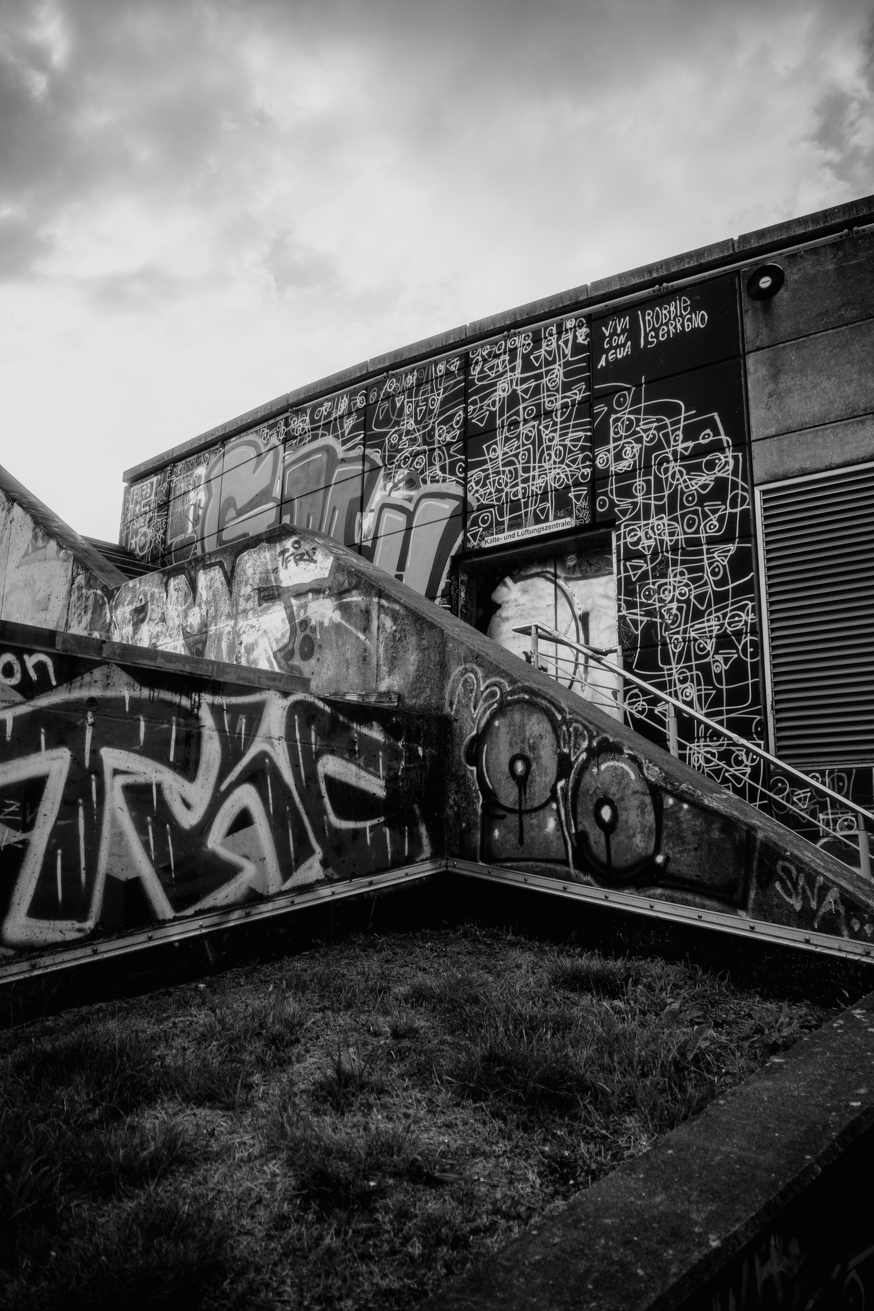 The black and white photo depicts a concrete building and stairway covered in various graffiti and street art, set against a cloudy sky.