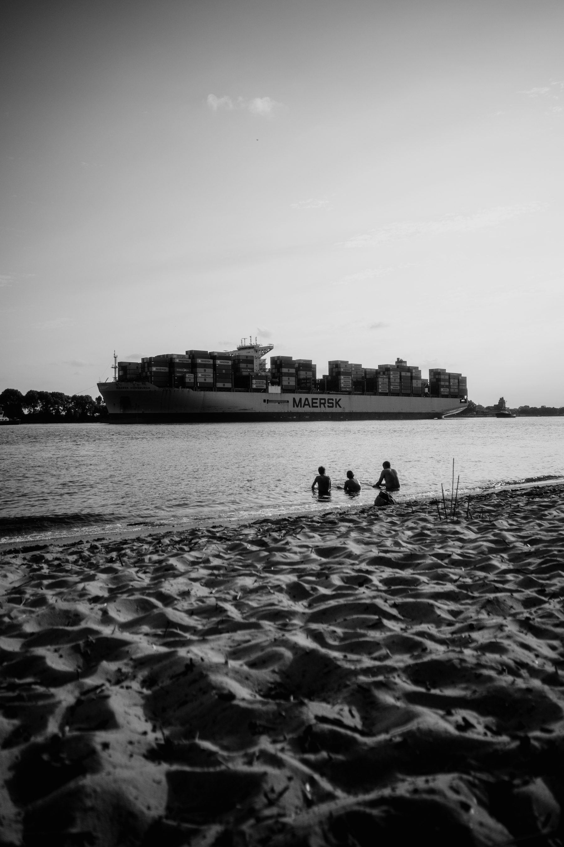 The image shows a large Maersk container ship passing by in the background while three people sit in shallow water near the shore, with sandy beach in the foreground, all captured in black and white.