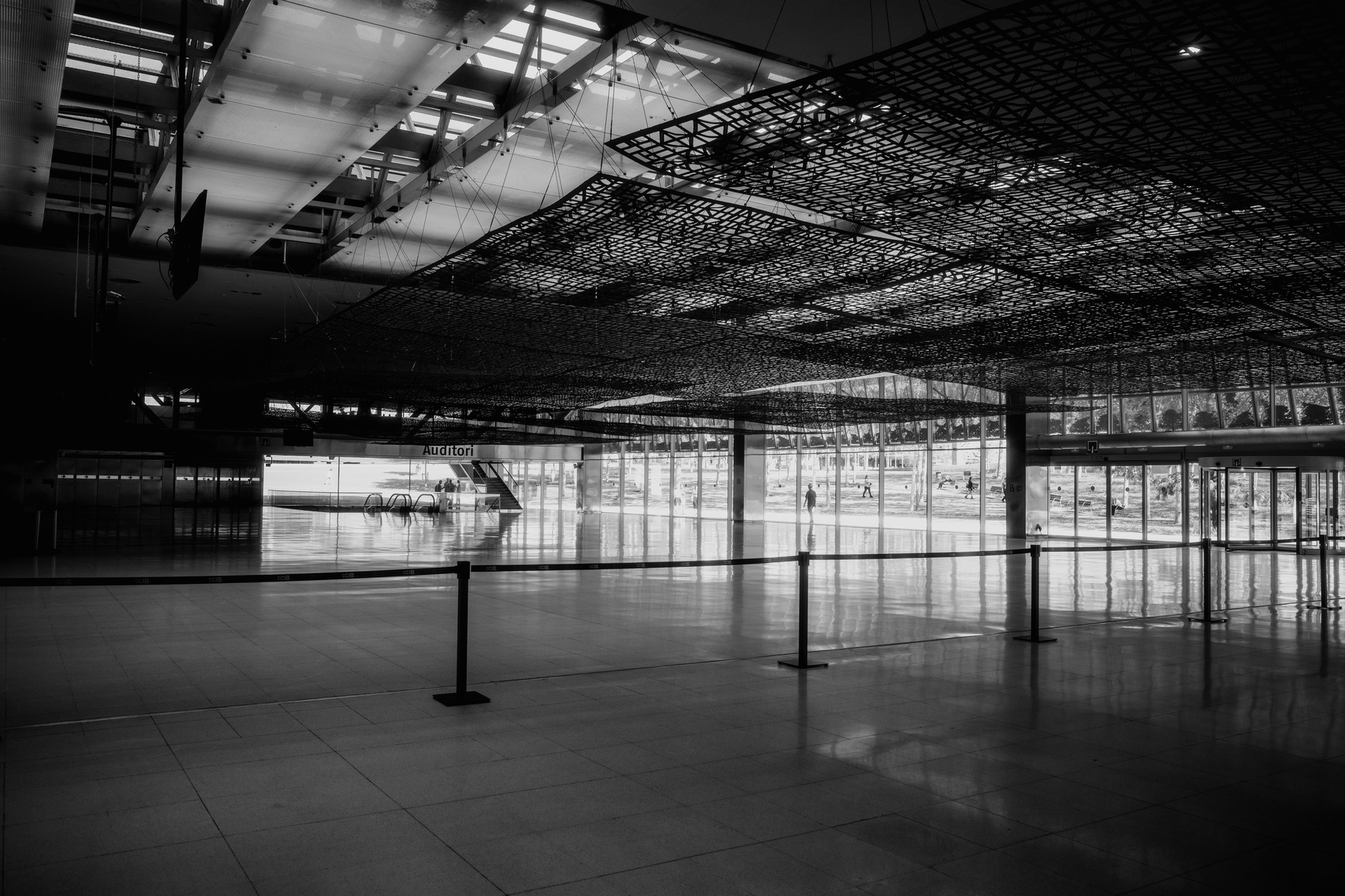 The image shows a spacious, modern interior with a high ceiling, metal lattice designs above, and a large glass window wall allowing light to flood in, with an empty hall leading to an “Auditori” and an escalator in the distance.