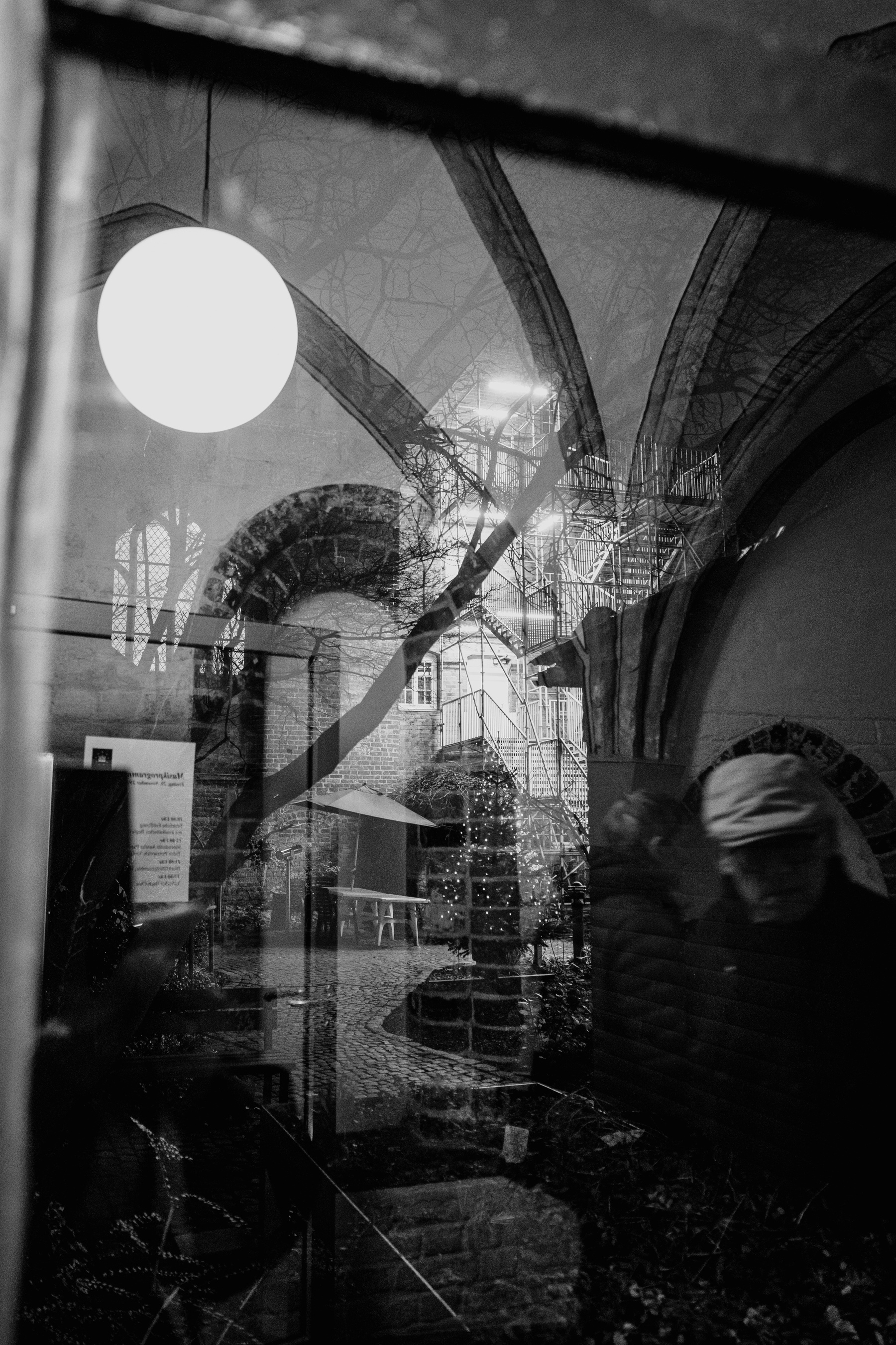 The black-and-white image captures a layered reflection of a gothic-style architecture, an illuminated stairwell, and cobblestone courtyard, blended with silhouettes of people in motion.