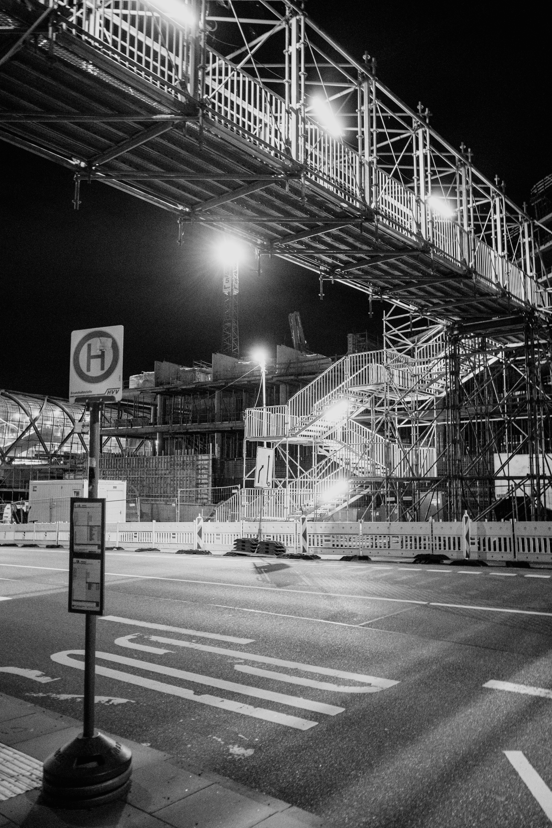 The image shows a nighttime construction site with a metal pedestrian bridge and staircase illuminated by bright lights, and a bus stop sign in the foreground.