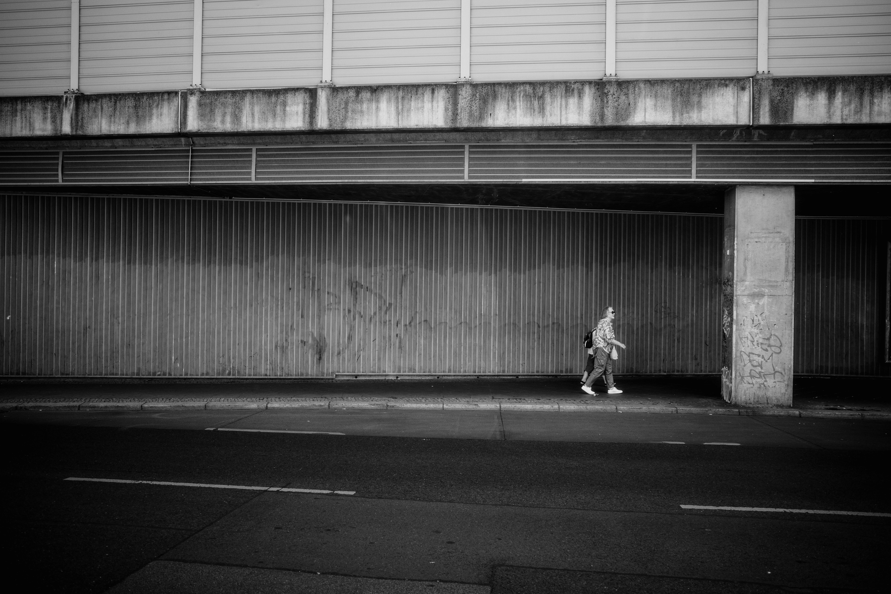 A person is walking alone along a deserted street under a concrete overpass, with the scene captured in black and white.