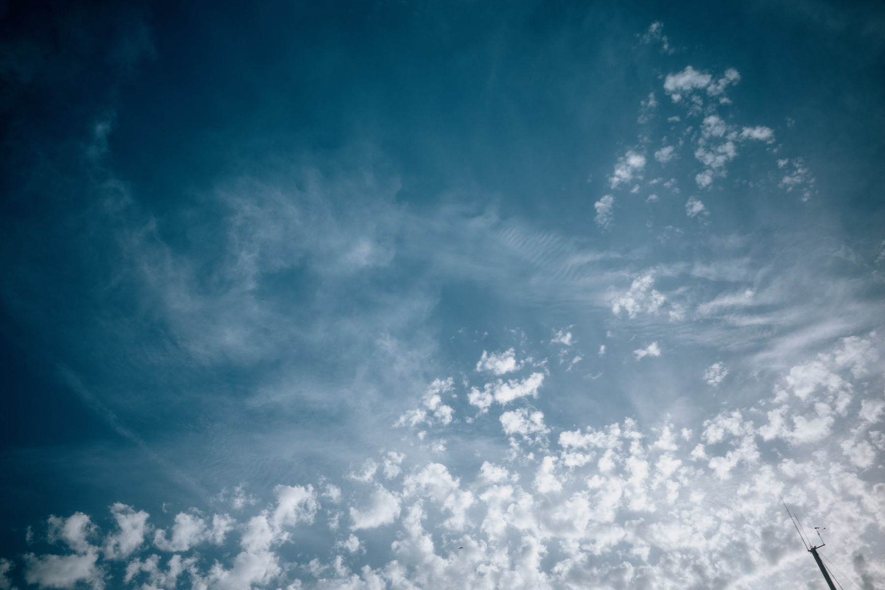 The image shows a blue sky with scattered clouds and a thin pole with a wind vane and anemometer in the lower right corner.