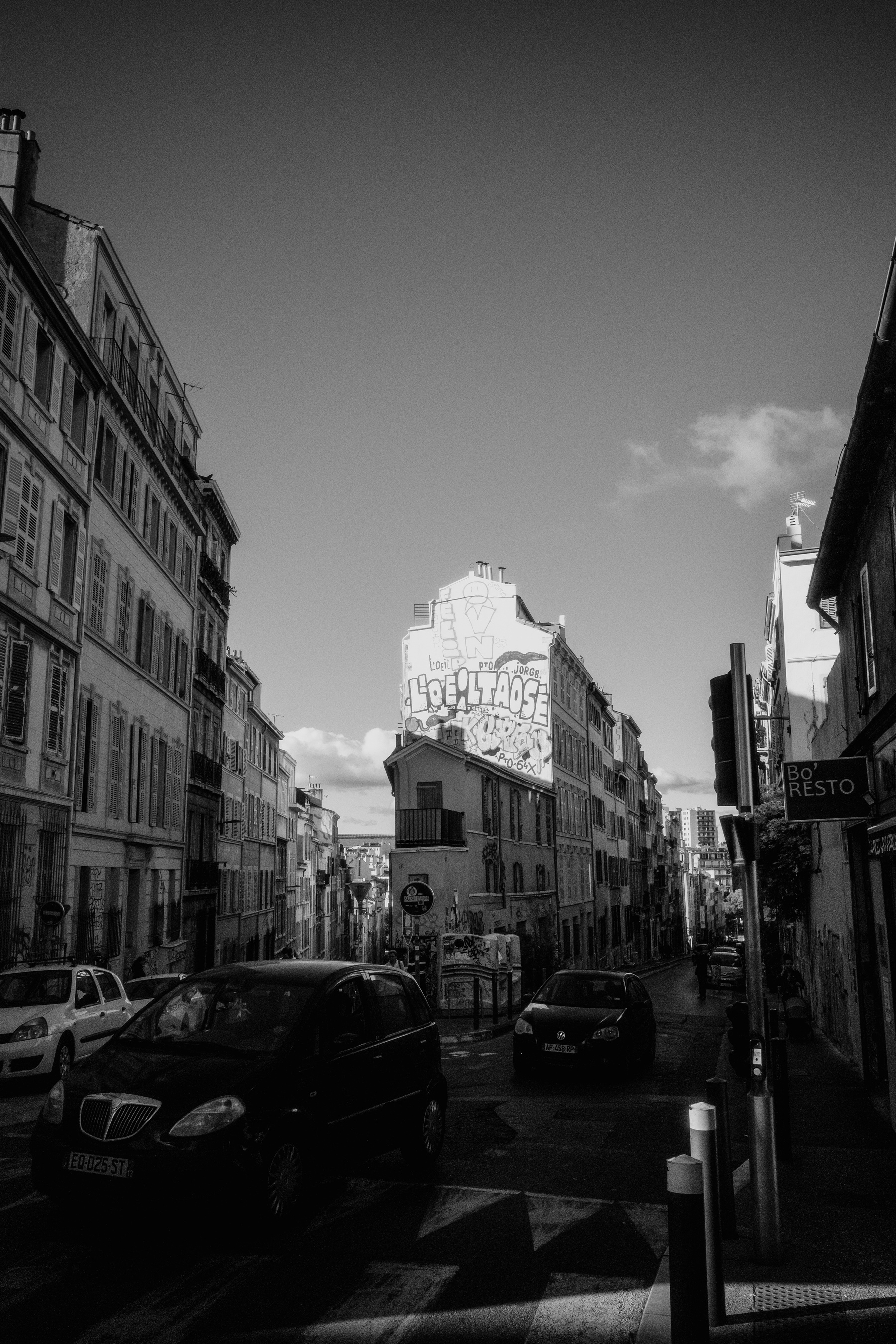 The image depicts a black-and-white urban street scene in Marseille with graffiti on a prominent building in the background, surrounded by older buildings and a few cars on the road.