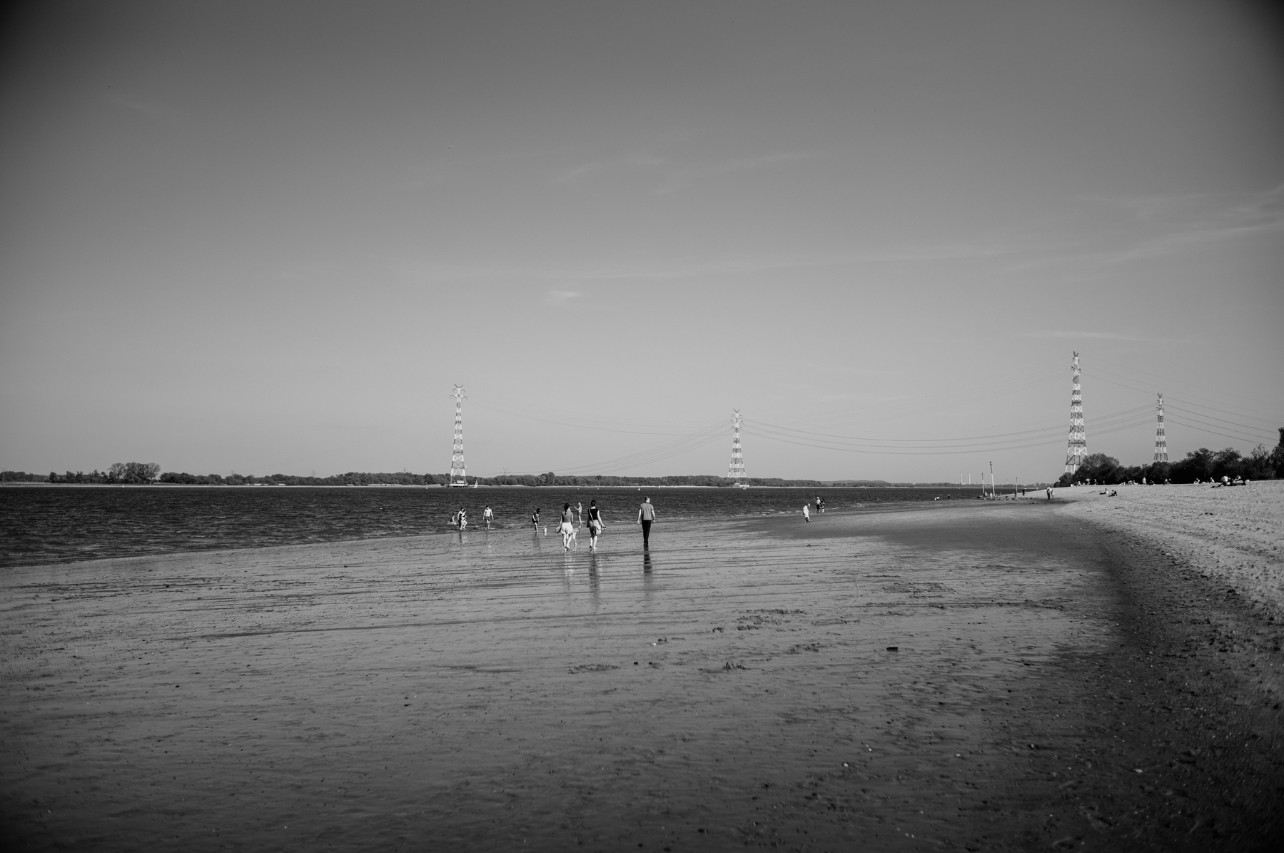 The image depicts people scattered along a wide, flat shore with shallow water, featuring large electricity pylons in the background under a clear sky, rendered in black and white.